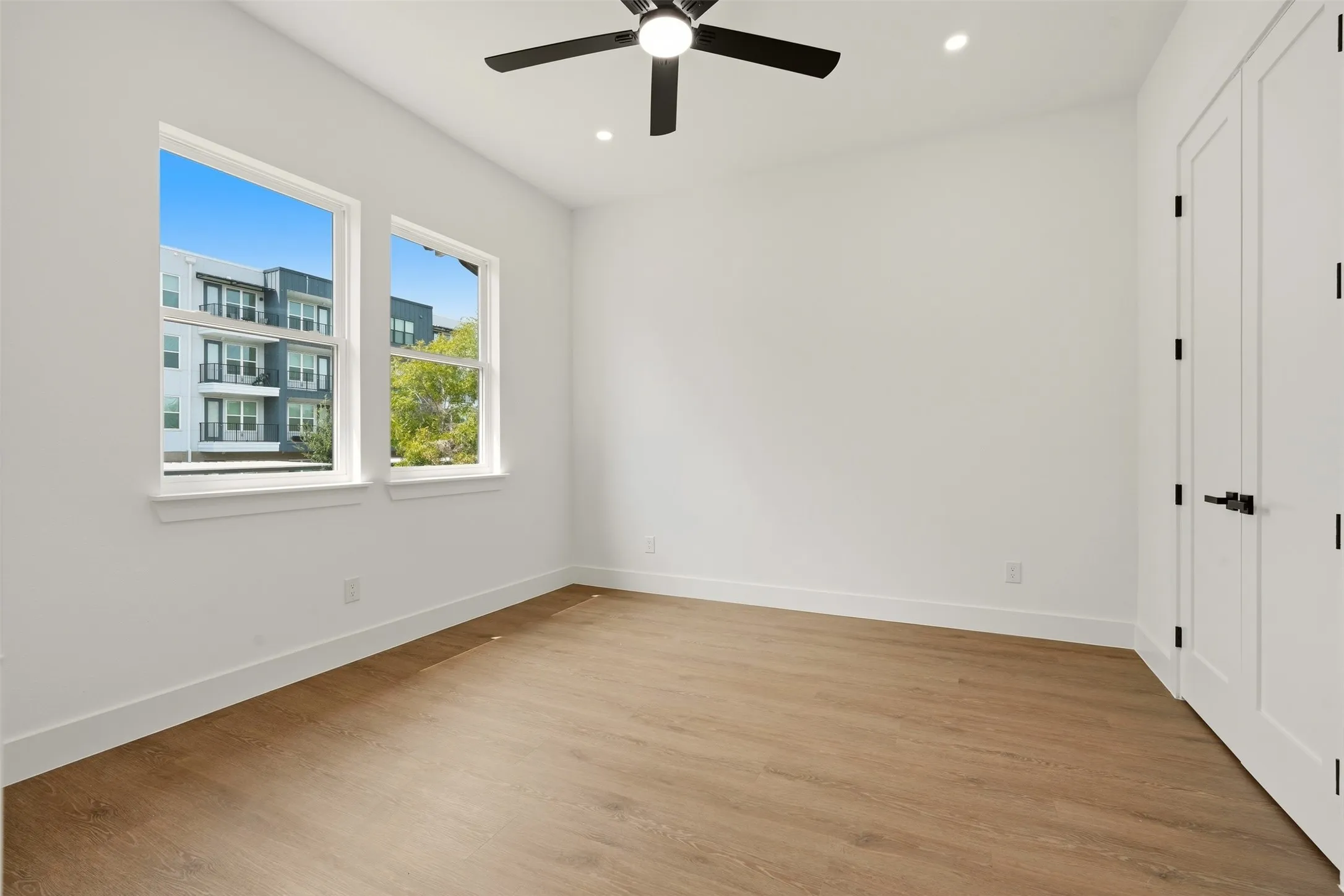 Two double-hung windows with white trim, wood-finish flooring, a contemporary ceiling fan, recessed lighting, and a paneled door with matte black hardware