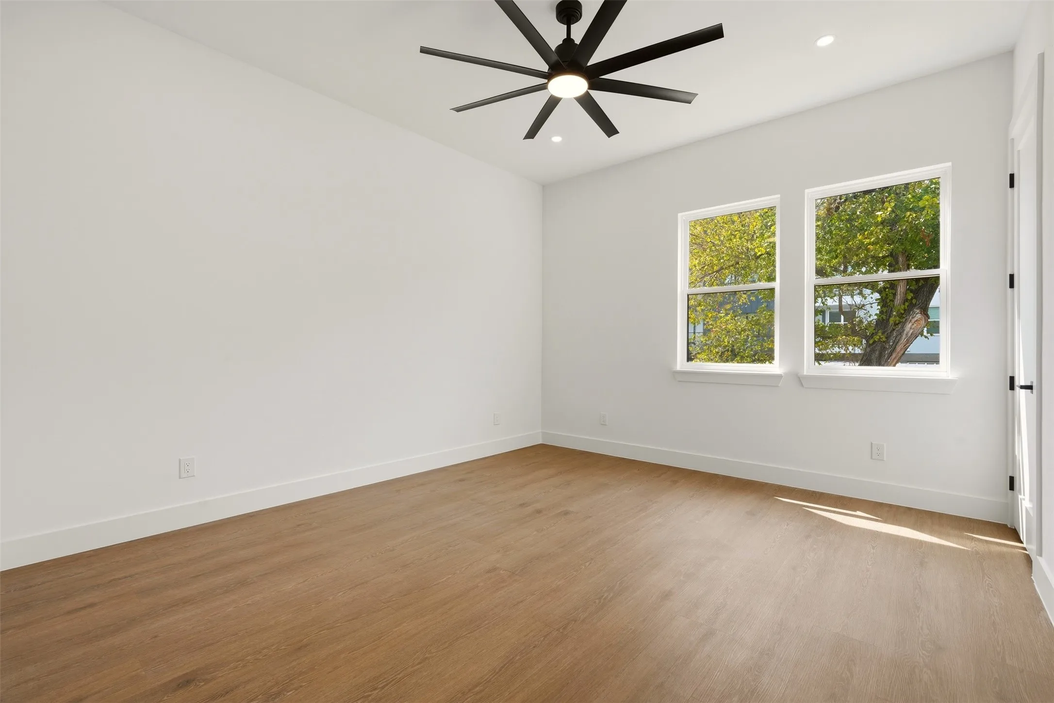 Spacious room featuring wood-finish flooring, two large windows, a modern ceiling fan, and recessed lighting