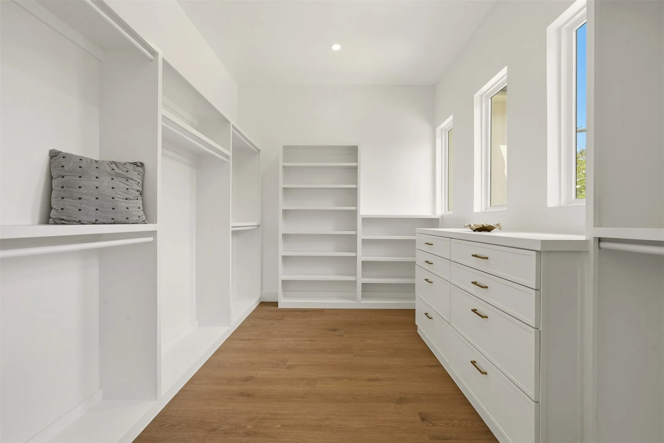 Spacious closet with wood-finish flooring, white built-in shelving, white built-in cabinetry with bronze hardware, and multiple windows