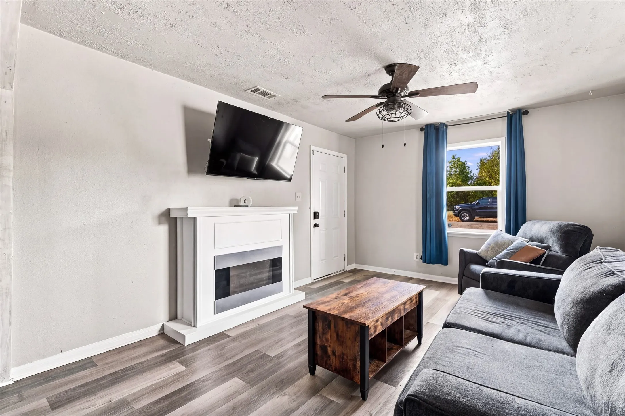 Wood-finish flooring, white fireplace surround, ceiling fan with light fixture, white door, and a prominent window