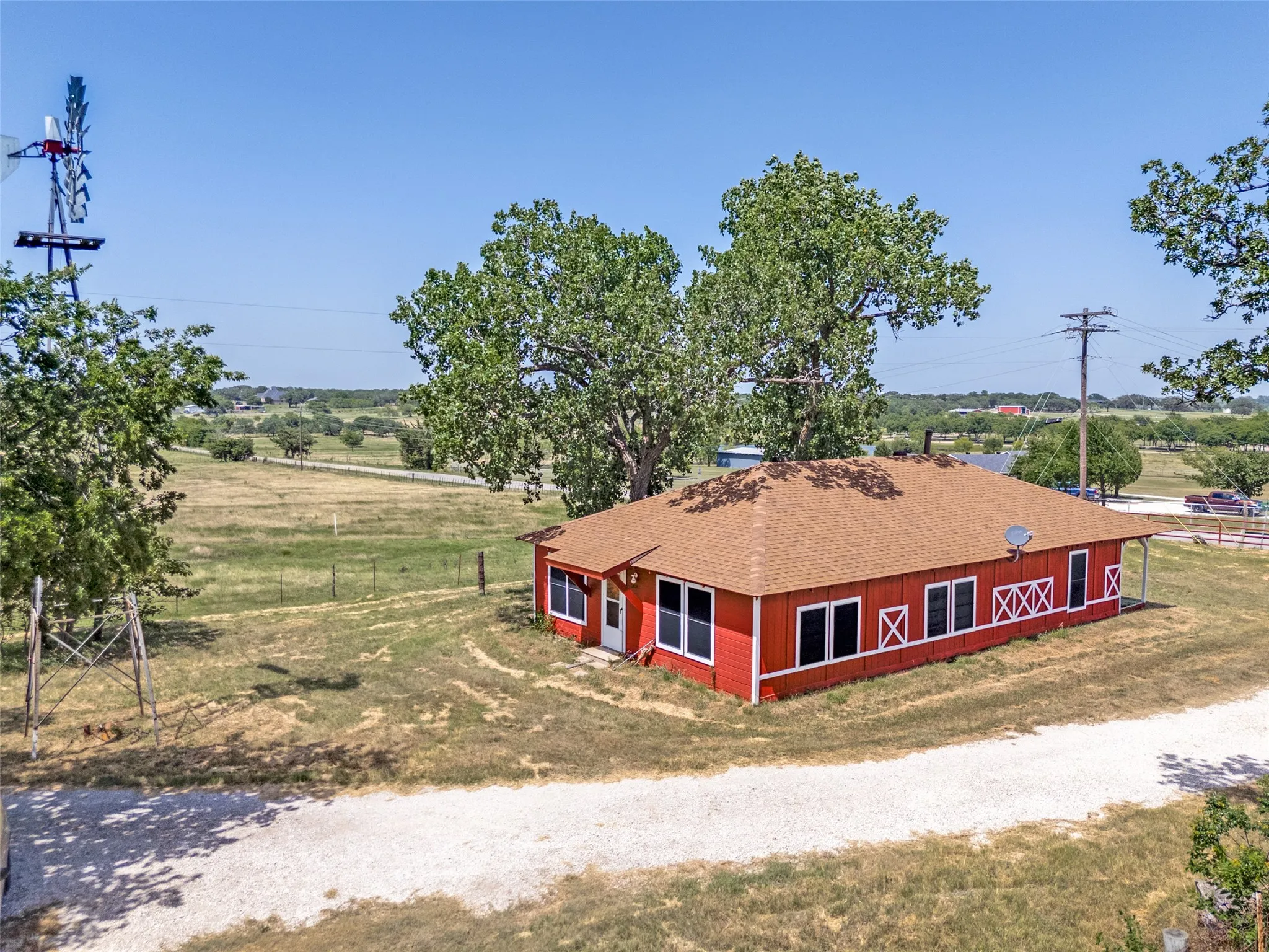 Red-sided structure featuring multiple windows, a tan shingle roof, and a gravel drive