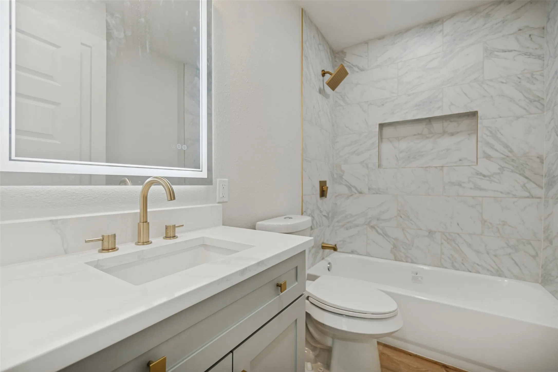 Integrated bathtub with a built-in shower niche and wall-mounted shower head, vanity with an undermount sink and gold-finish fixtures, and a back-lit mirror