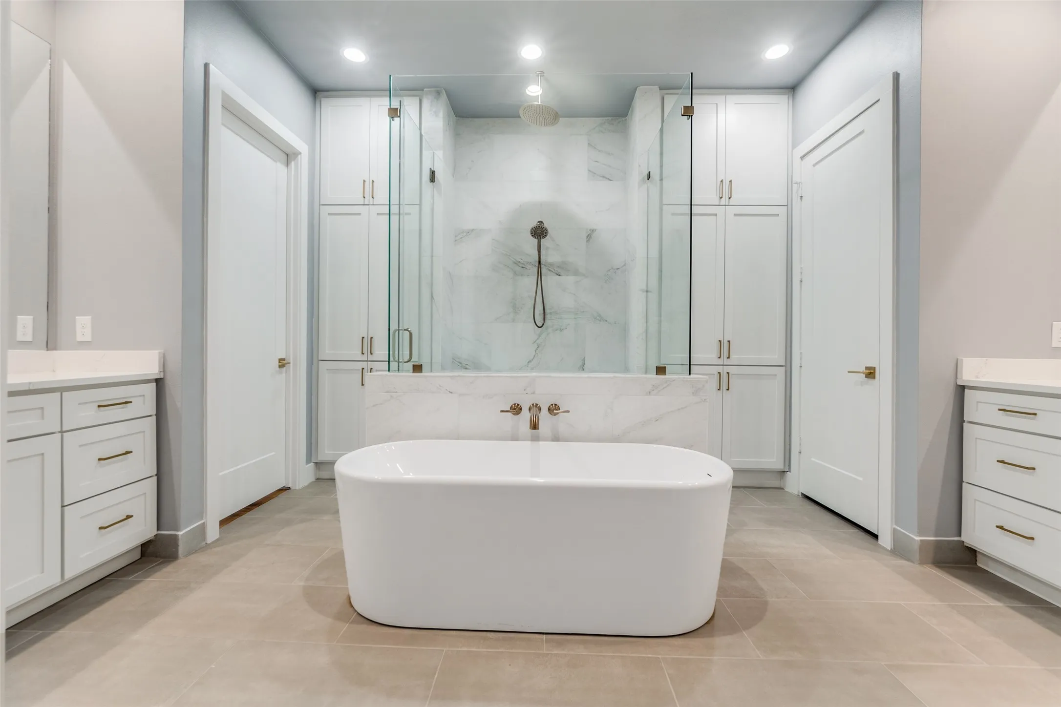 Freestanding soaking tub, dual vanities with stone countertops, glass-enclosed shower, built-in cabinetry, and tile flooring