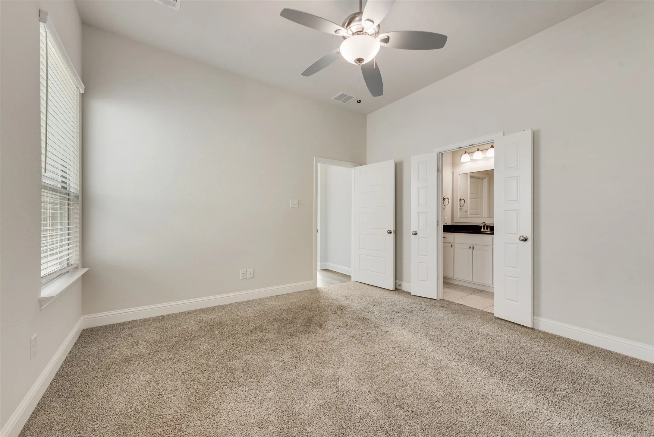 Vaulted ceiling, ceiling fan with integrated lighting, carpeted flooring, window with blinds, and an attached bathroom featuring a vanity with a dark countertop