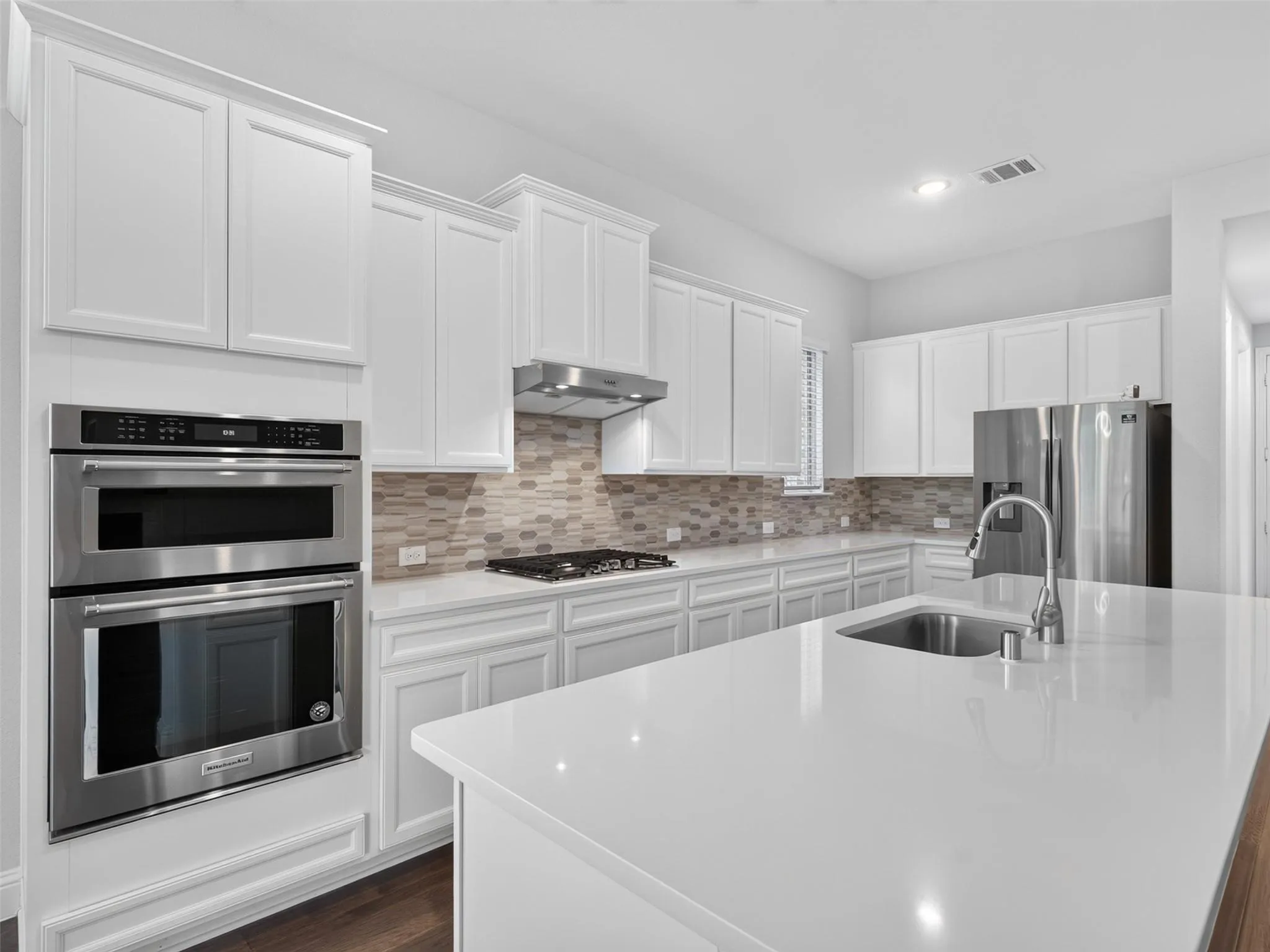 White shaker-style cabinetry, stainless steel appliances, light-toned tiled backsplash, white countertop surfaces, and a central island with an undermount sink