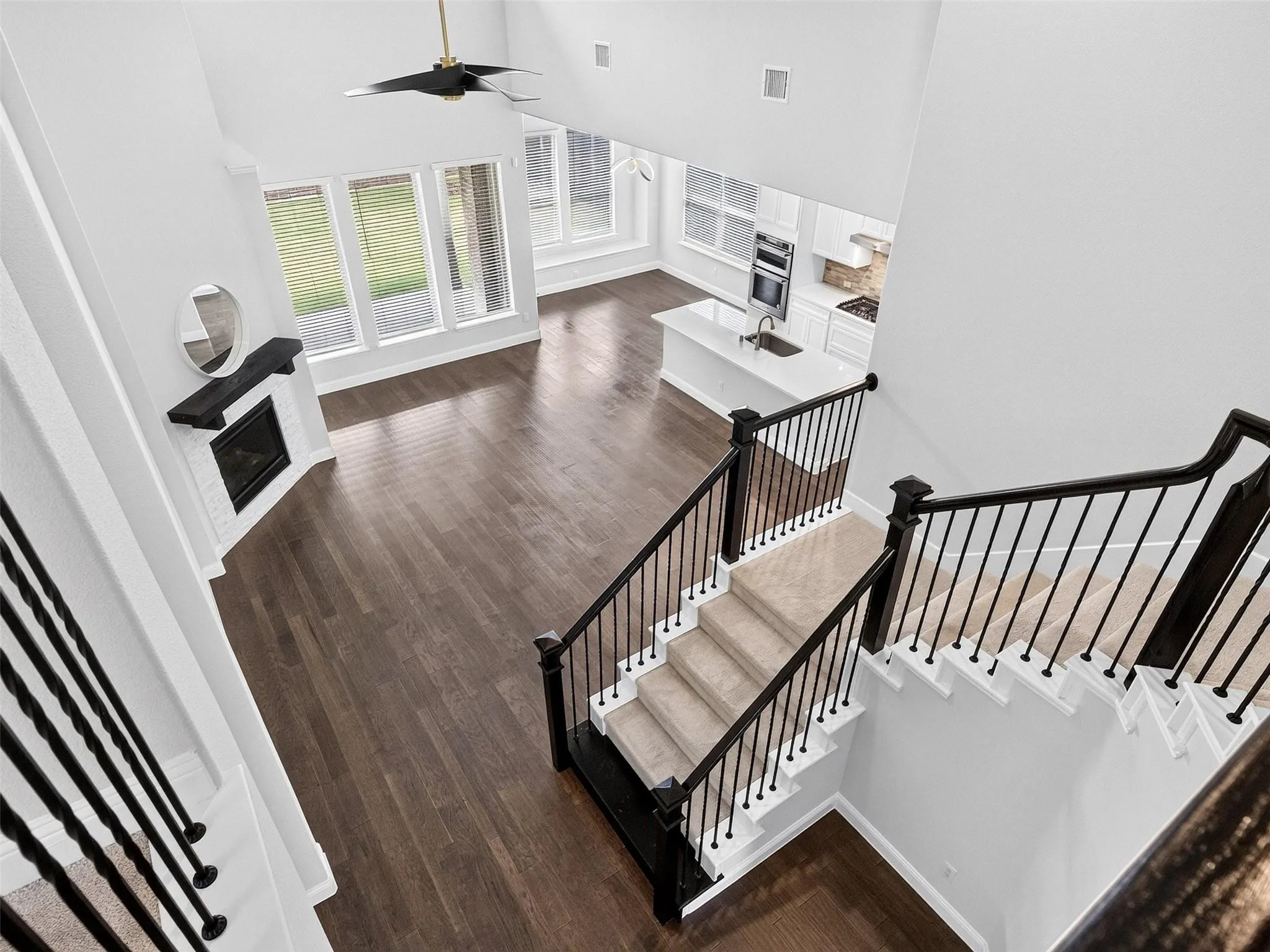 Vaulted ceiling with a ceiling fan, wood-finish flooring, white brick fireplace with a dark mantel, white kitchen island with a sink, and a carpeted staircase with dark railings