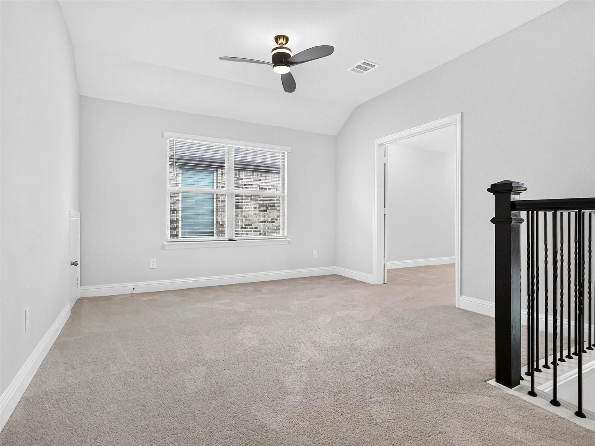 Neutral-toned carpeting, ample natural light from a large window, a ceiling fan, and a black wrought iron newel post and baluster