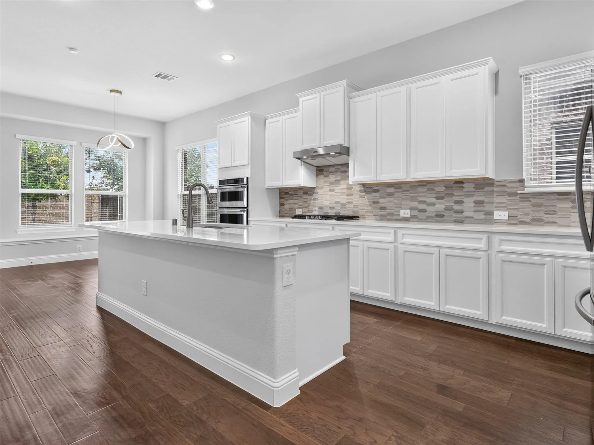 Expansive kitchen island with a quartz countertop and integrated sink, stainless steel appliances, white cabinetry, decorative tile backsplash, and wood-finish flooring