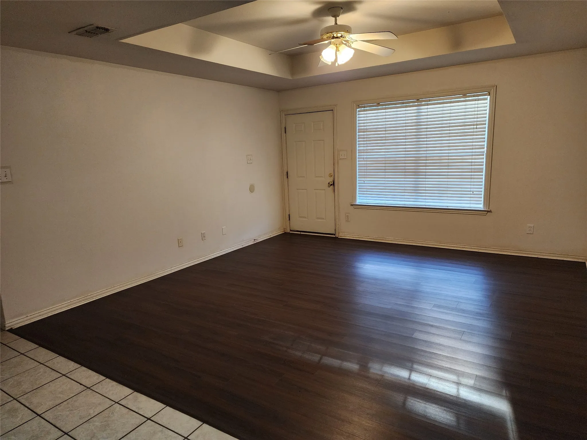 Wood-finish flooring, tray ceiling, ceiling fan with integrated lighting, window with blinds, and a white entry door