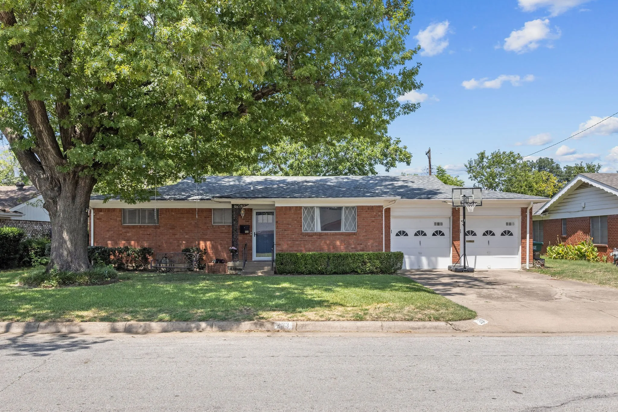 Brick exterior featuring a two-car garage with arched panel doors, mature front yard tree, established shrubbery, and a concrete driveway