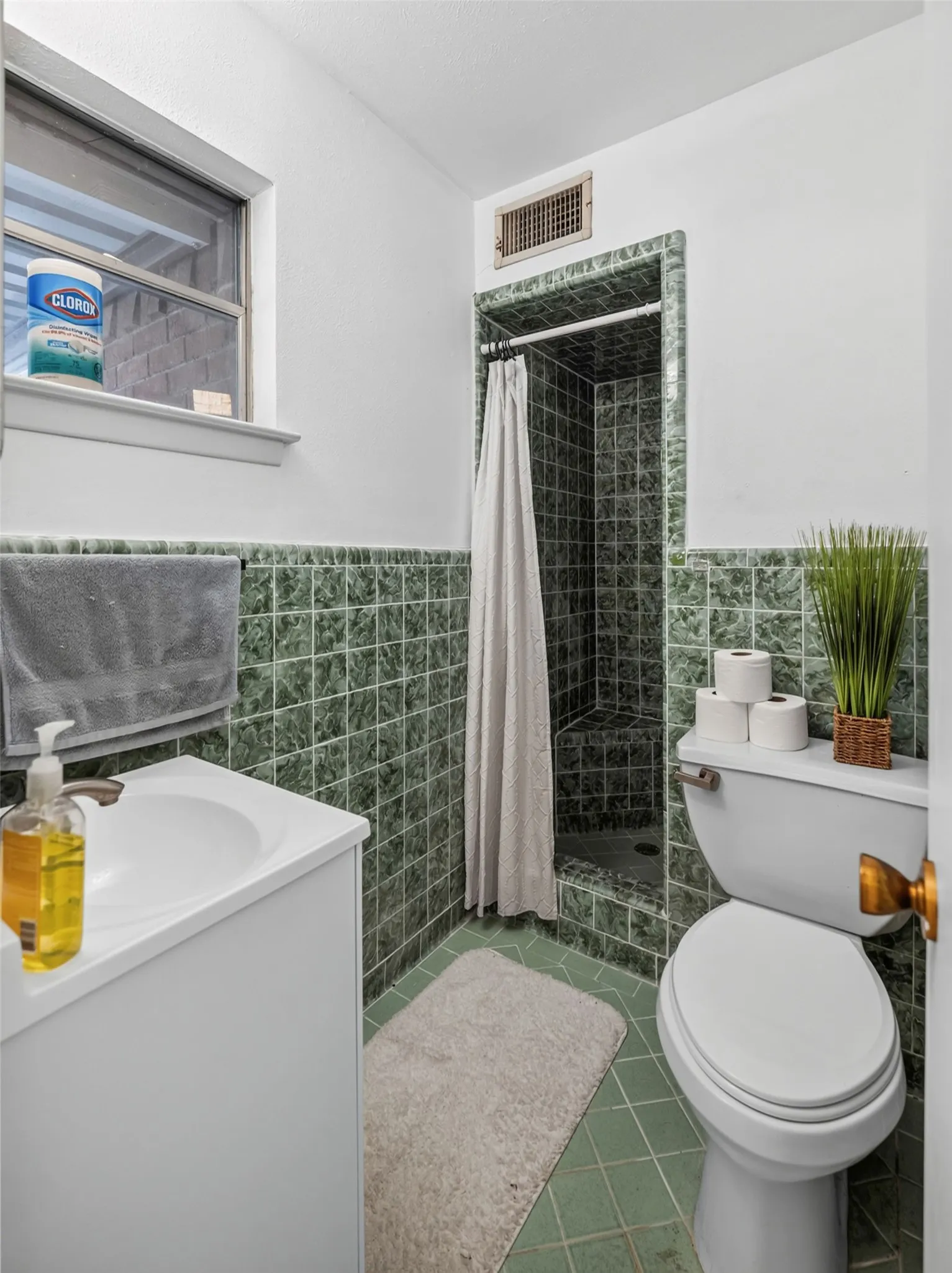 Compact bathroom featuring a shower with green subway tile surround, coordinating green floor tiles, a white vanity with an integrated sink, a white toilet, and a window with a sill