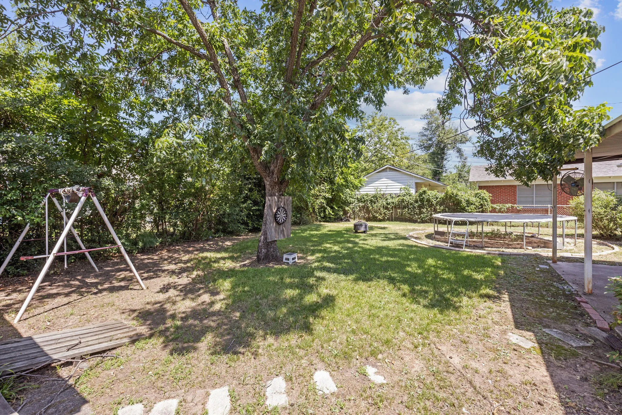 Brick exterior featuring a two-car garage with arched panel doors, mature front yard tree, established shrubbery, and a concrete driveway