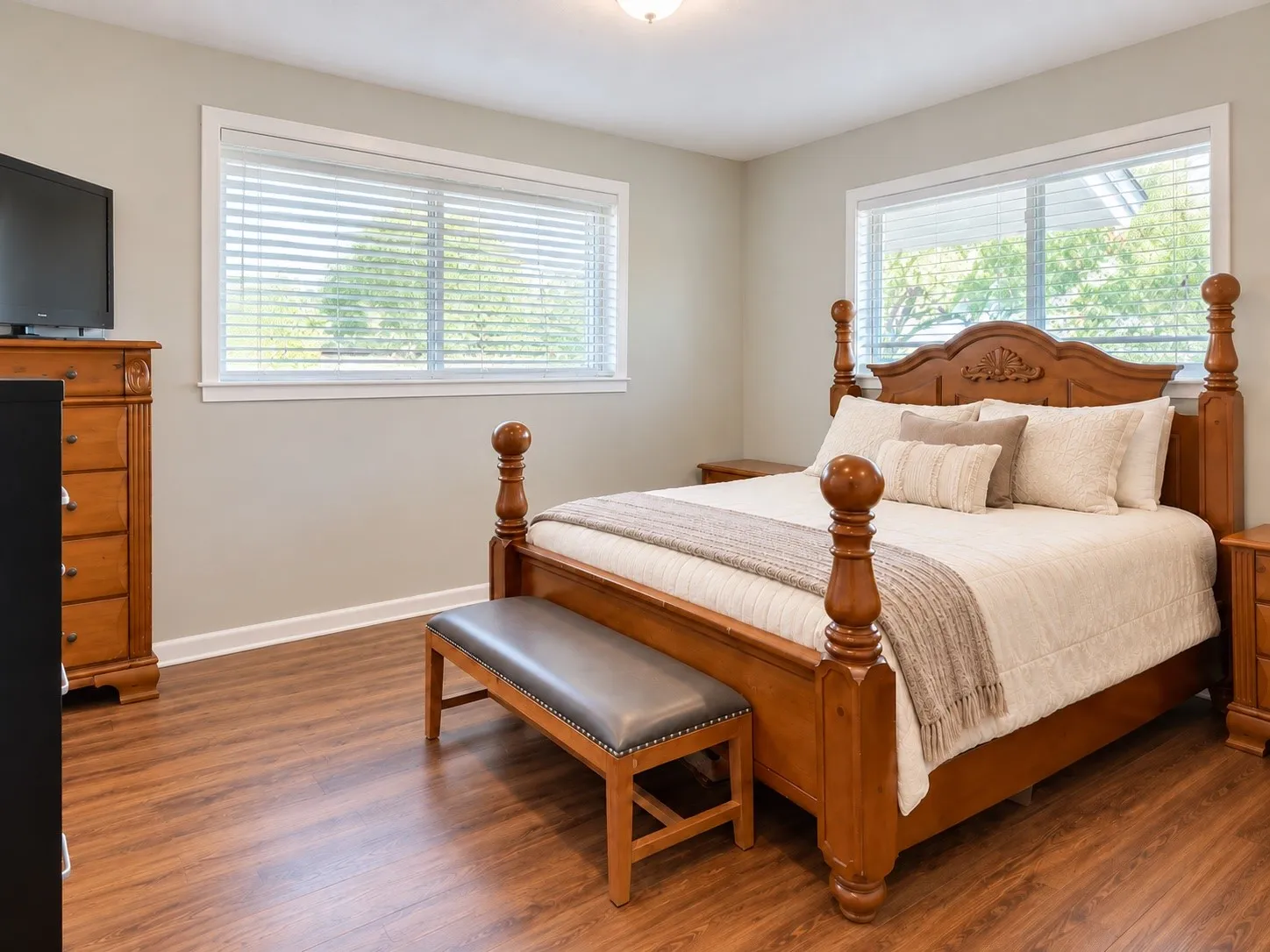 Spacious primary bedroom featuring a four-poster wood bed frame, hardwood flooring throughout, abundant natural light from dual casement windows with plantation shutters, and a dedicated dresser with drawers