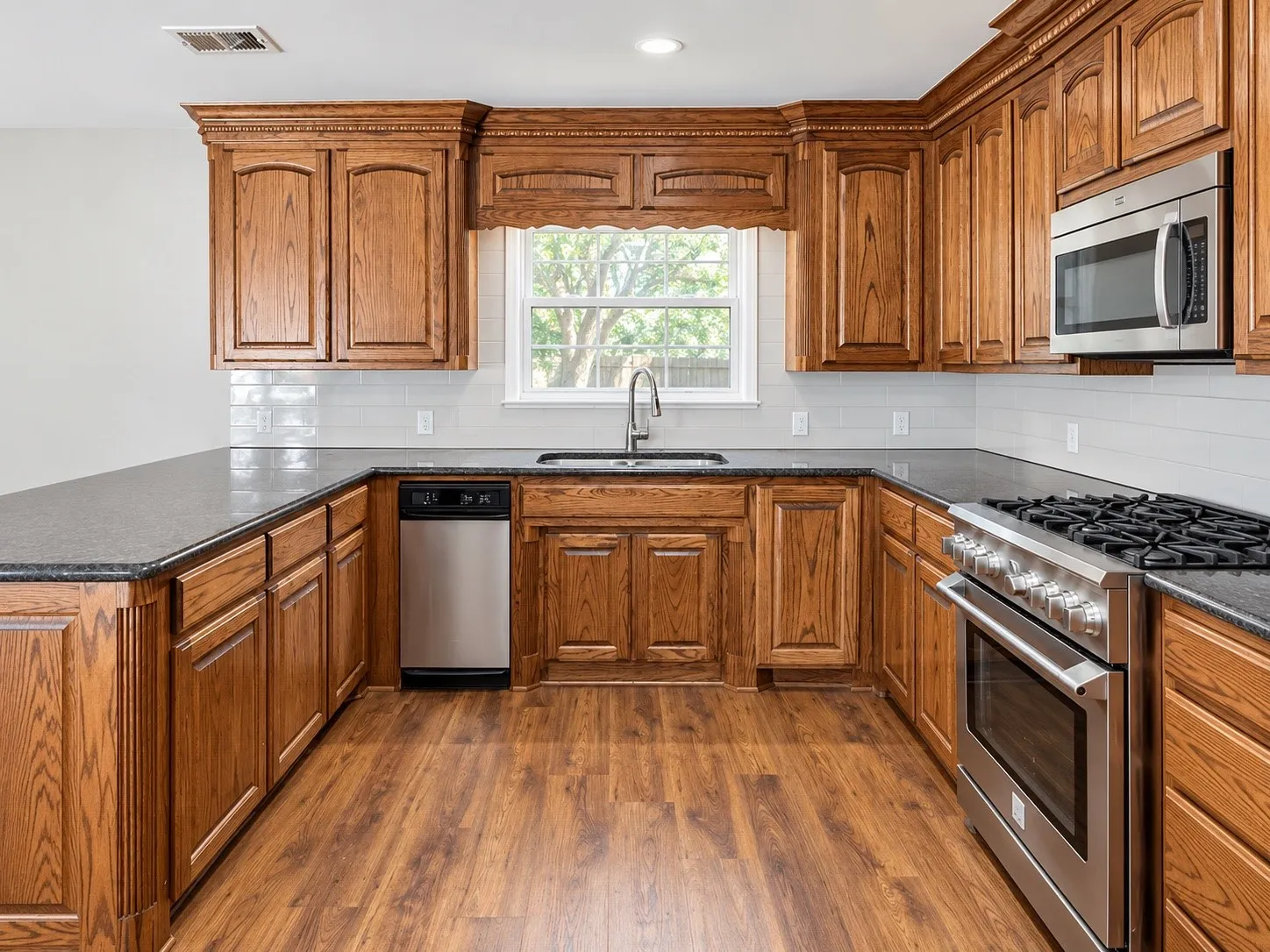 U-shaped kitchen with wood-finish flooring, dark countertops, wood cabinetry, stainless steel appliances, and a white subway tile backsplash