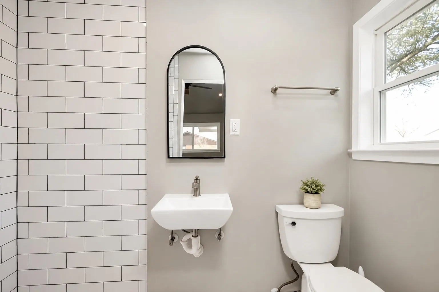 White subway tile surround, wall-mounted sink with chrome faucet, arched mirror with black frame, toilet, and window with white trim
