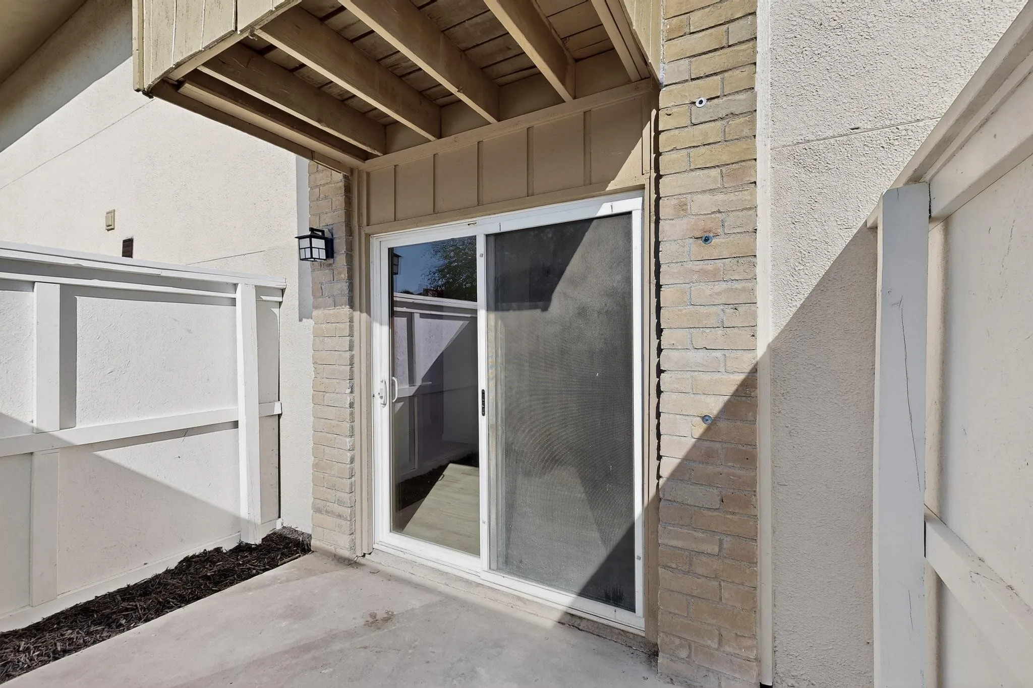 Private patio featuring a concrete surface, sliding glass door, brick facade, wood siding, and an exterior light fixture