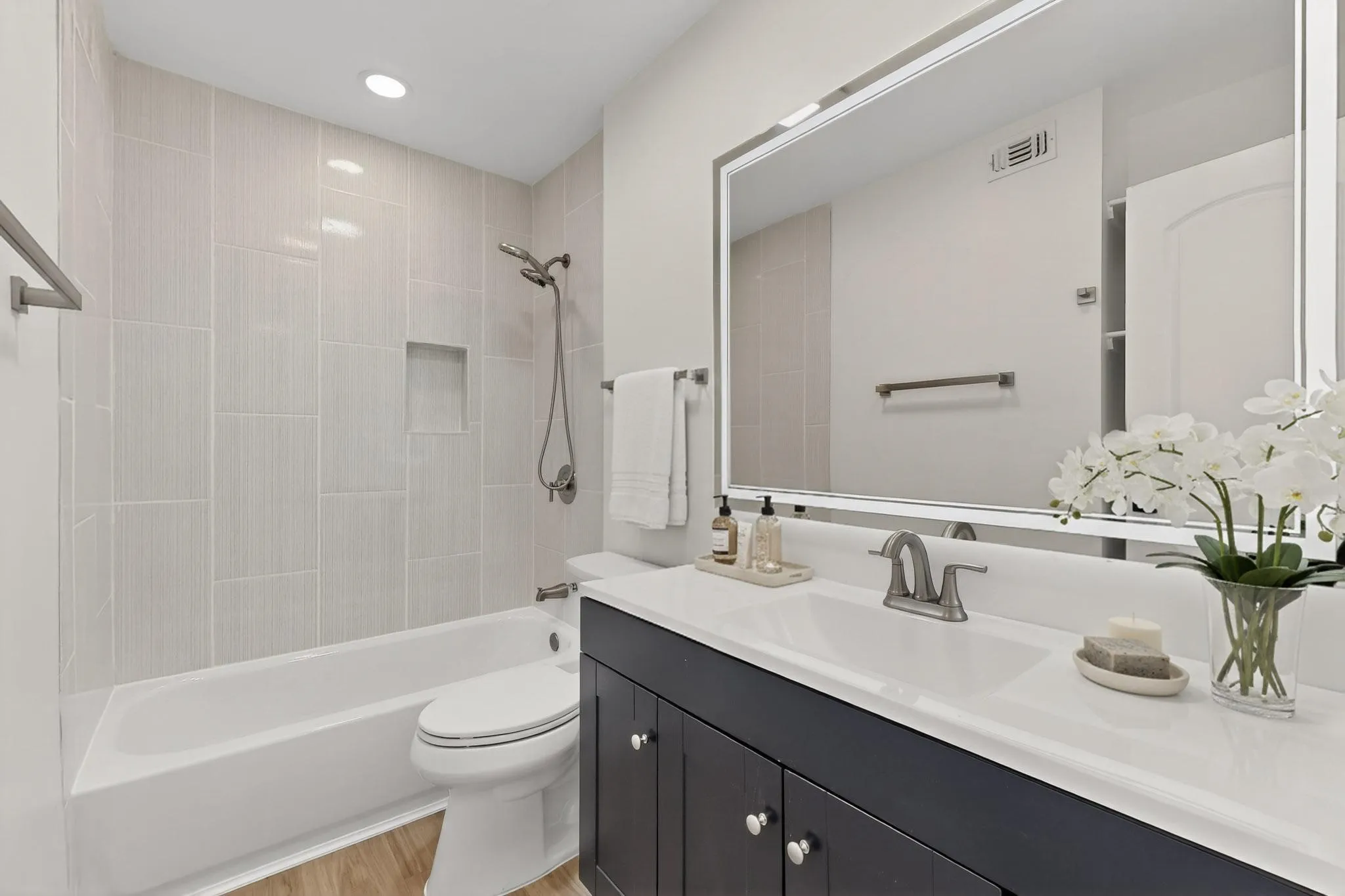 White ceramic bathtub with a shower niche, light-toned vertical tile surround, wood-finish flooring, a dark-finish vanity with a white countertop, and a large wall-mounted mirror