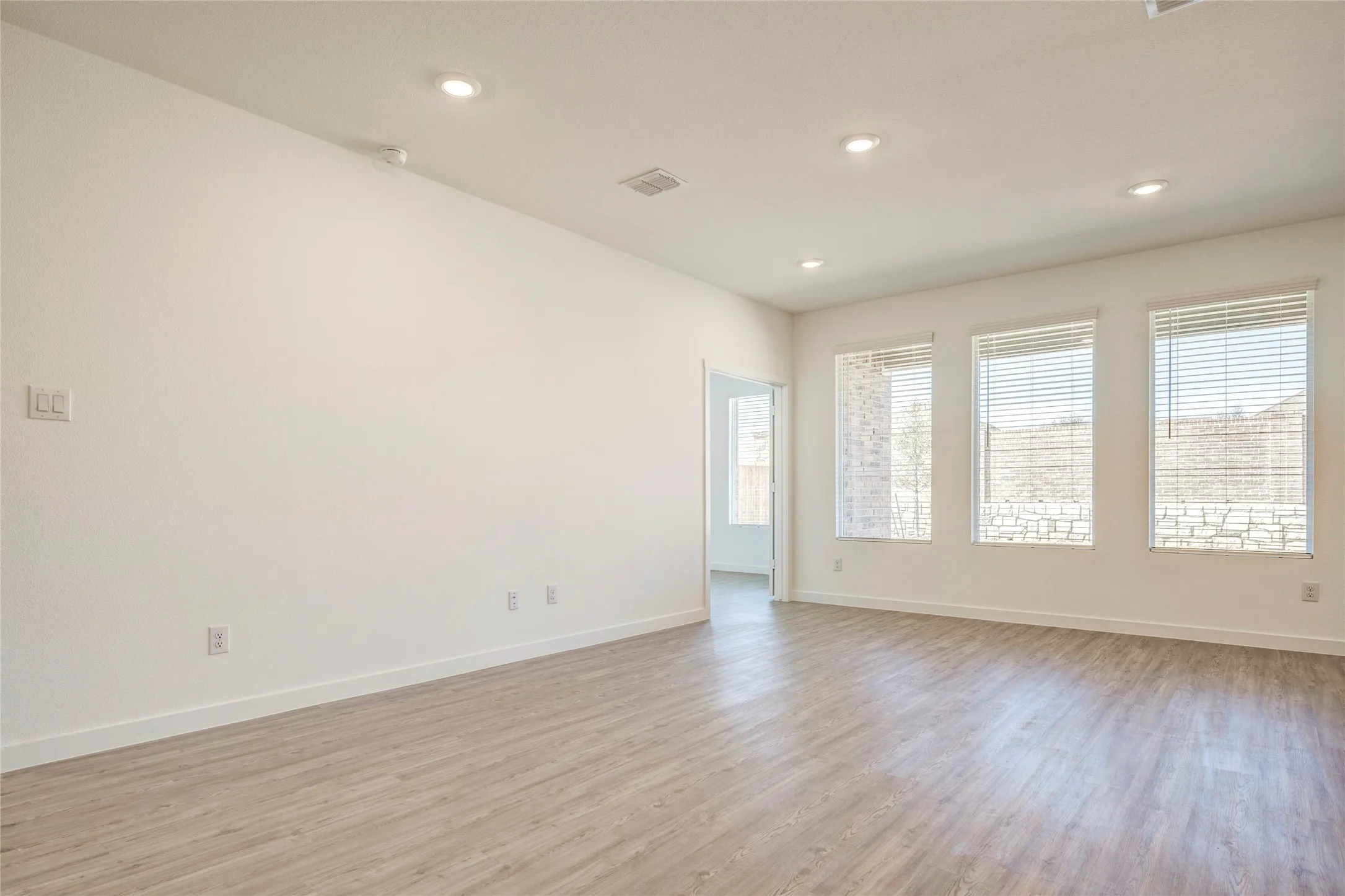 Sunny living room with recessed ceiling lighting, multiple windows with blinds, light LVP flooring, and white painted walls...