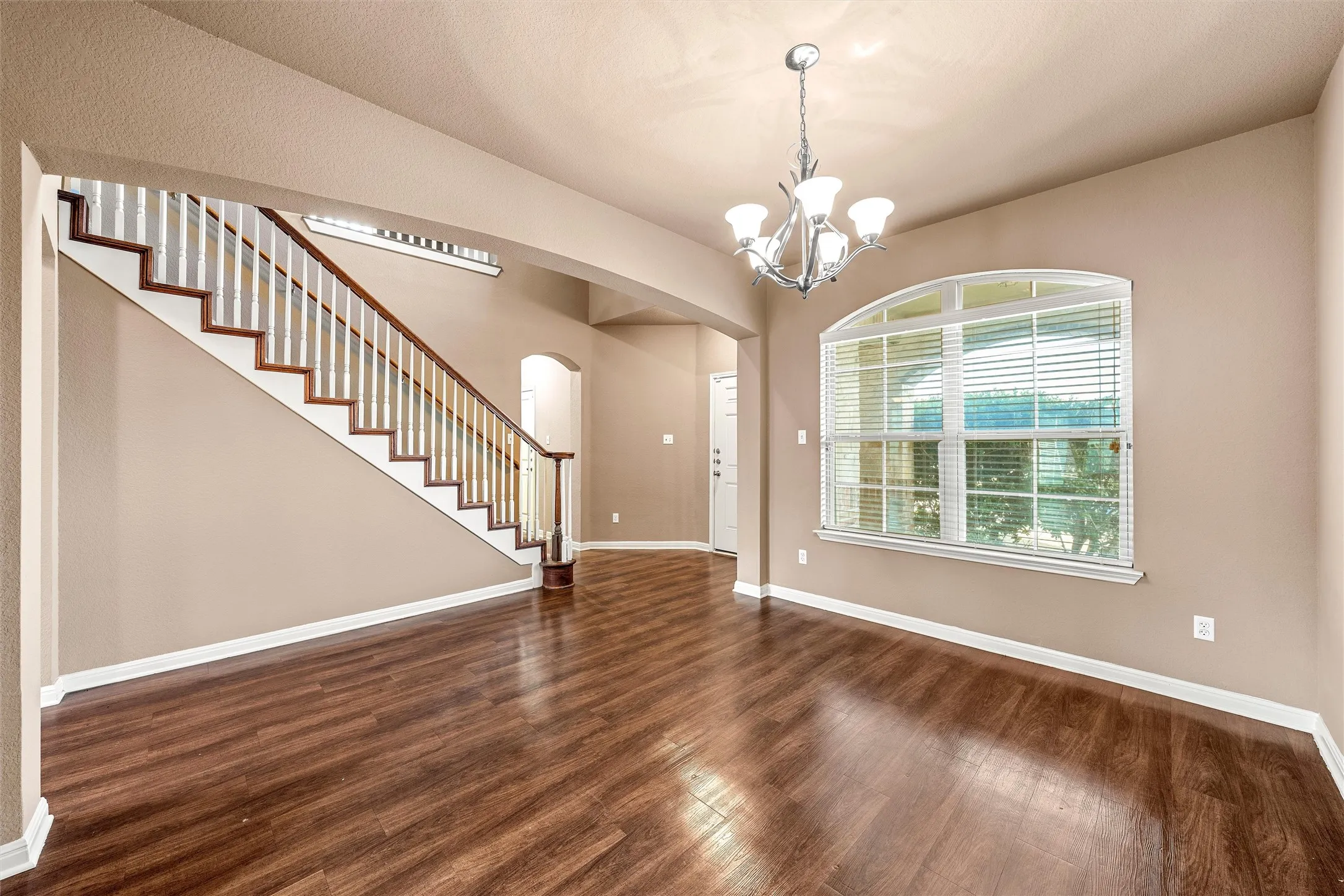Arched window with blinds, dark wood-finish flooring, staircase with white risers and wood handrail, polished chrome chandelier, arched doorway