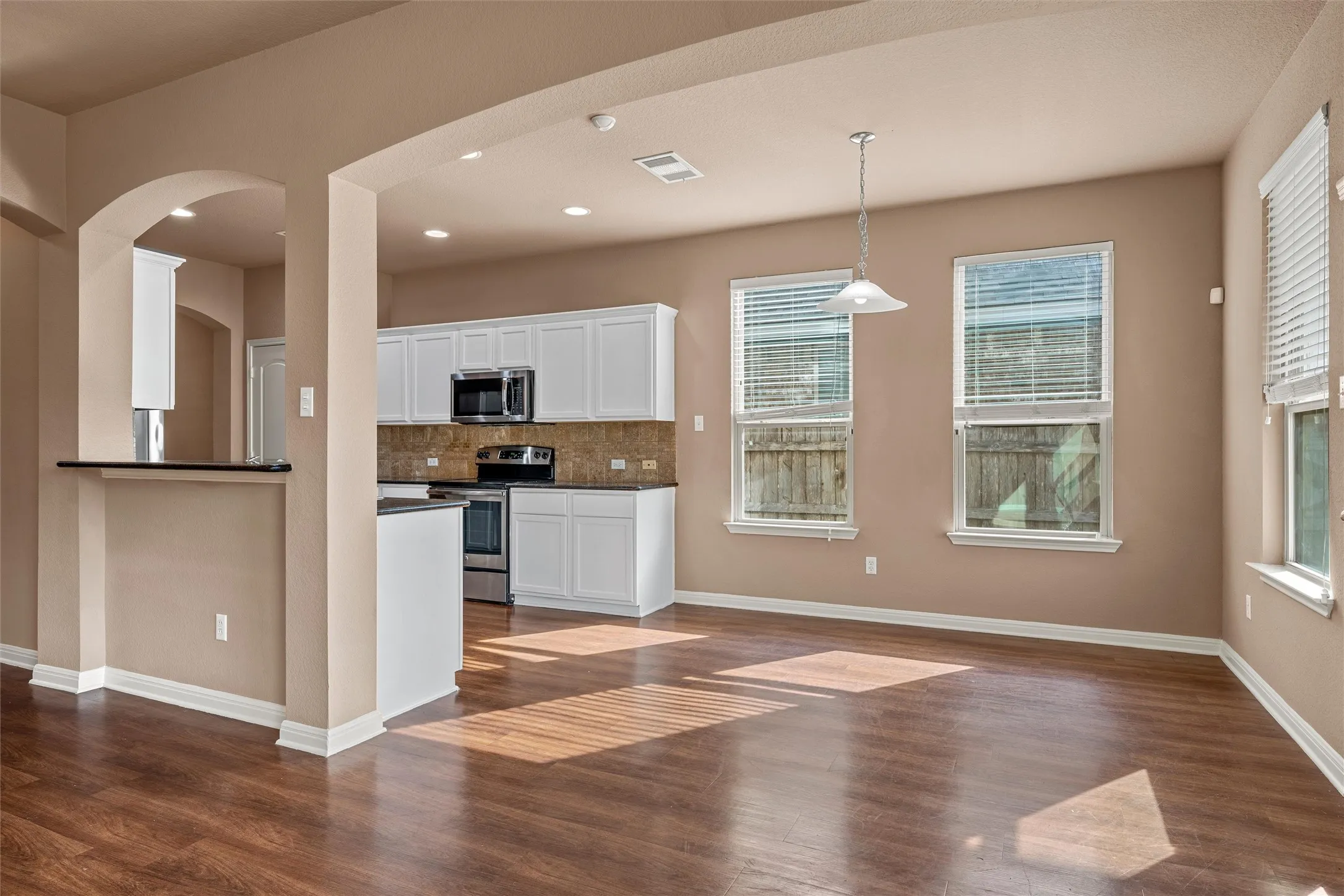 Open-concept living space featuring wood-finish flooring, white cabinetry, integrated stainless steel appliances, and multiple windows with blinds