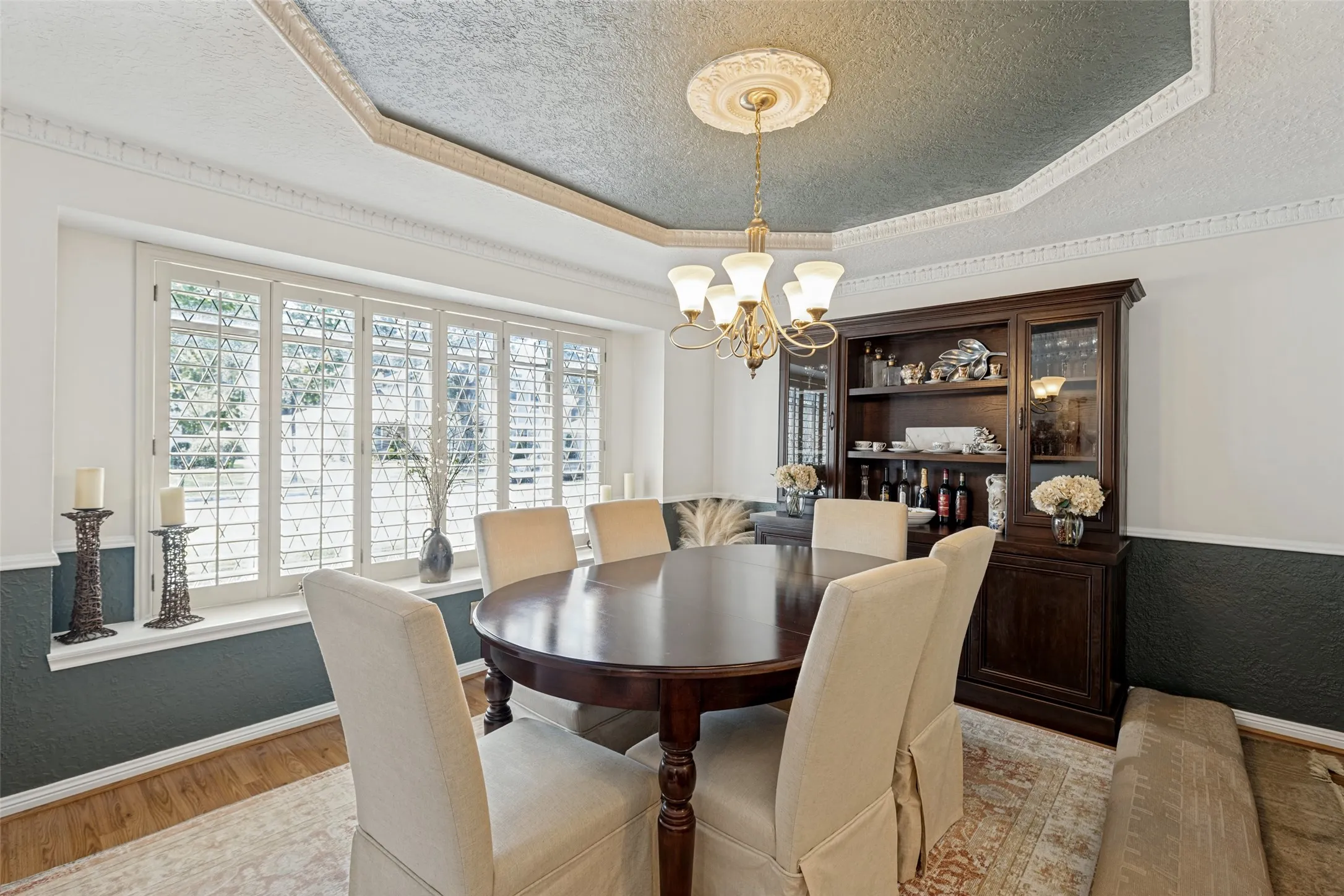 Tray ceiling with decorative molding, chandelier, bay window with plantation shutters, wood-finish flooring, and built-in display cabinetry