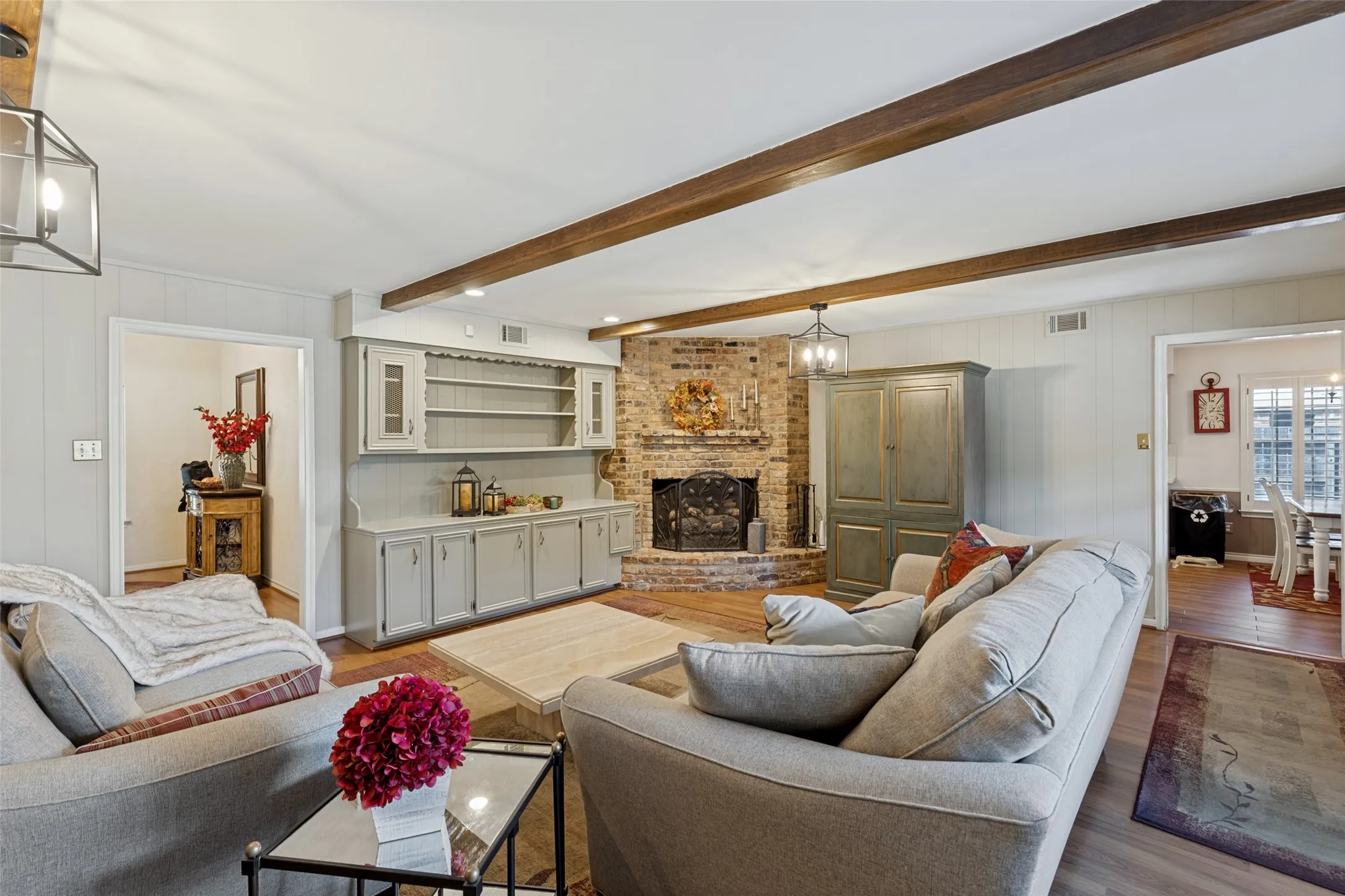 Exposed wood beams, wood-finish flooring, brick fireplace, built-in cabinetry with open shelving, and recessed lighting