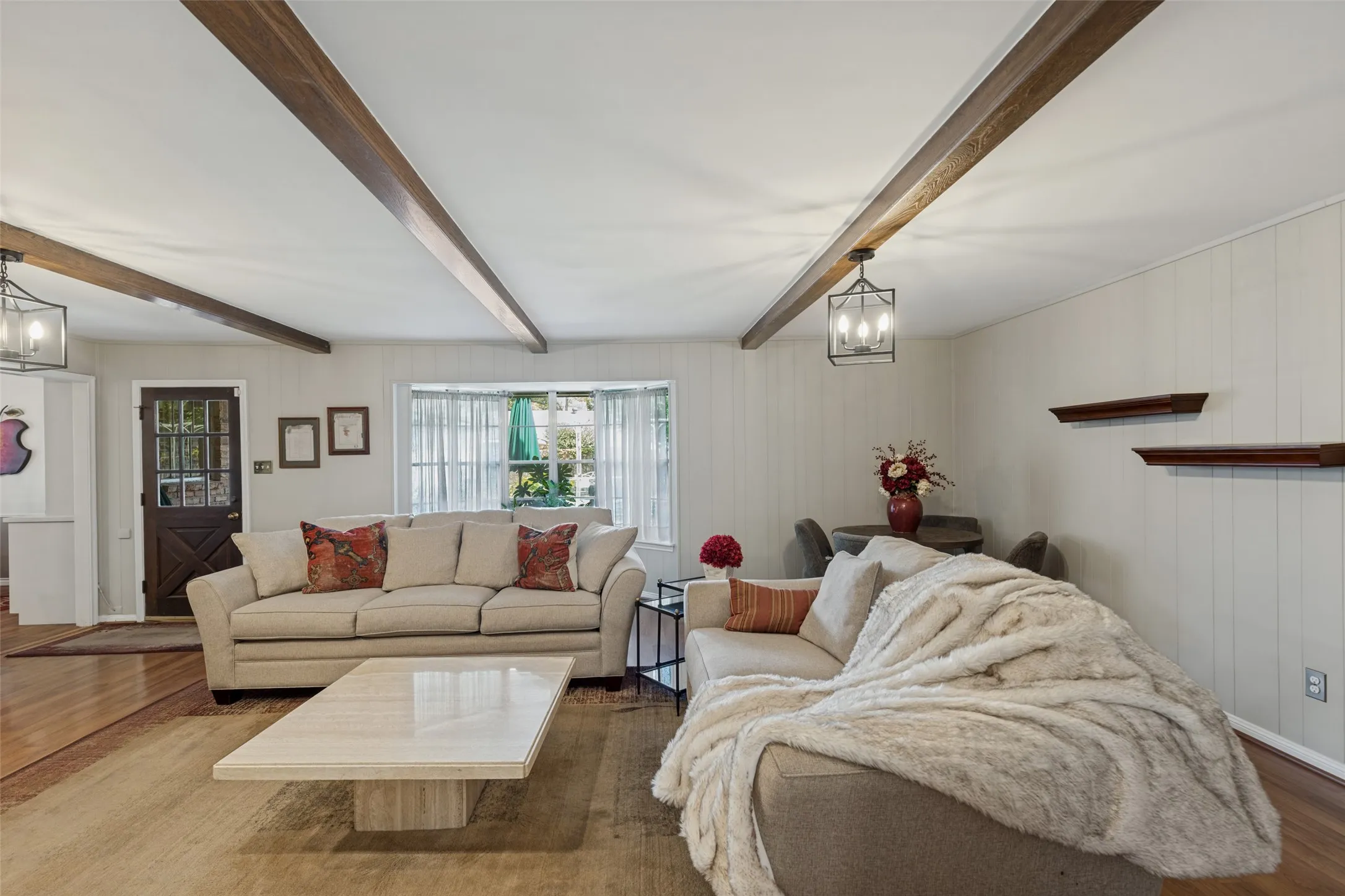 Wood-finish ceiling beams, wood-finish flooring, white paneling, and a large window with sheer curtains