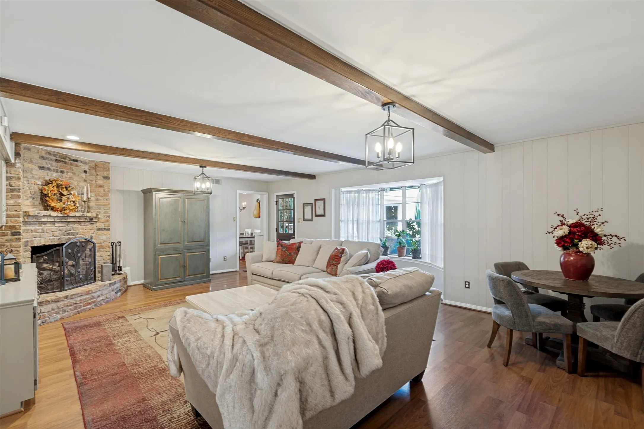 Exposed wood beam ceiling, brick fireplace with hearth, wood-finish flooring, bay window, and paneled wall accents