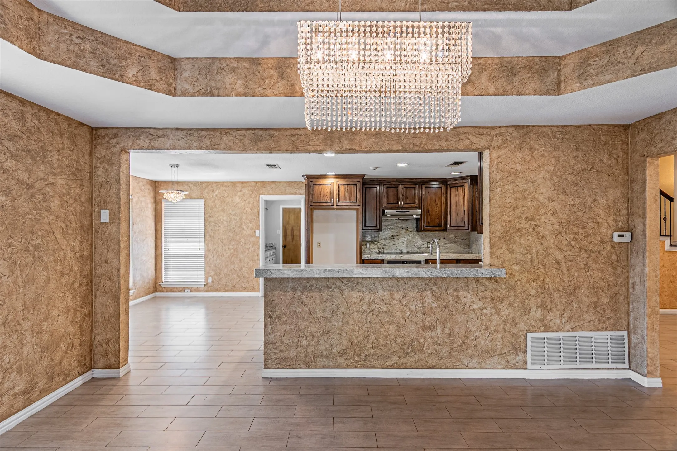 Recessed ceiling with an ornamental chandelier, tile flooring, and a kitchen pass-through with a breakfast bar