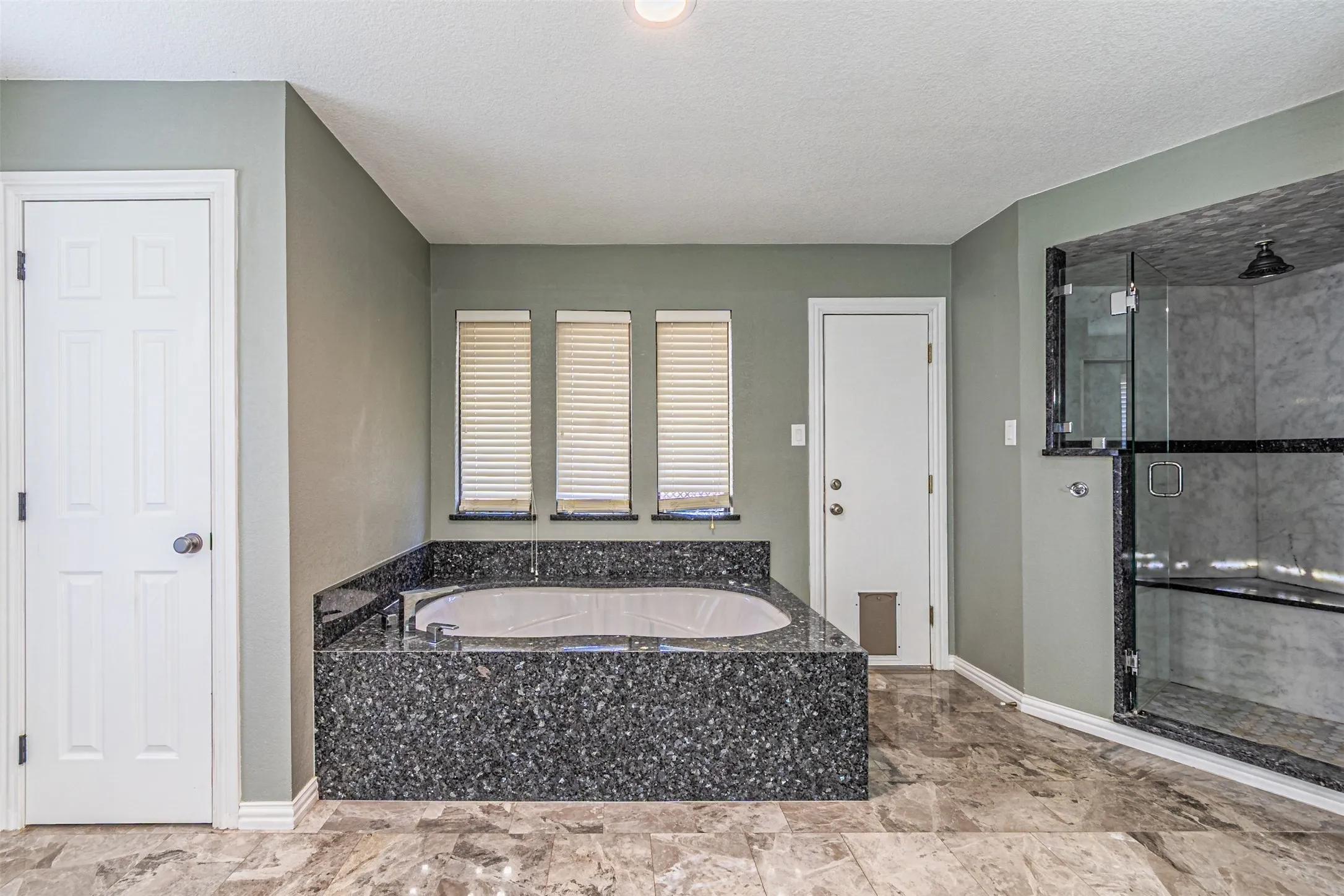 Soaking tub with a dark stone surround, three windows with blinds, glass-enclosed shower with a dark stone base, and tiled flooring
