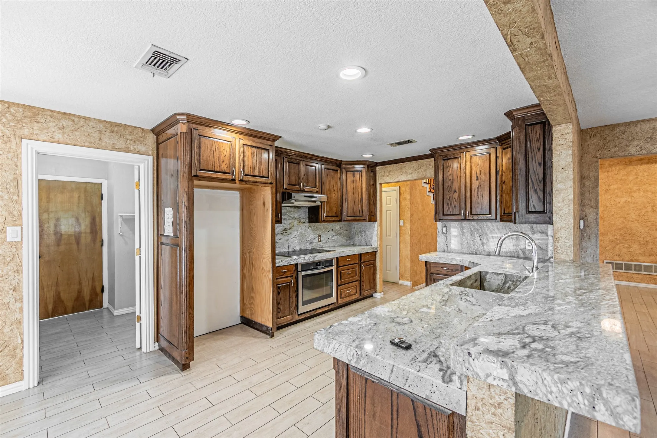 Kitchen featuring wood cabinetry, light-toned stone countertops, a stainless steel sink with a gooseneck faucet, an integrated cooktop, and light-toned tile flooring