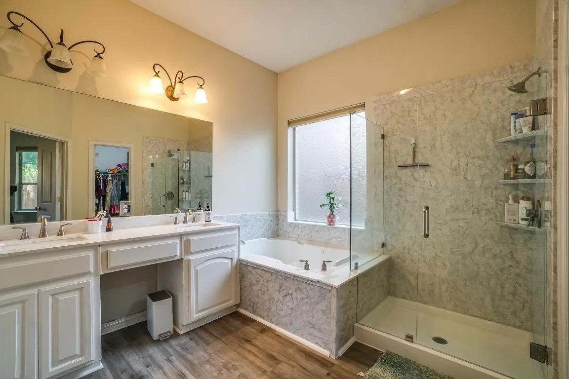 Dual vanity with undermount sinks, wood-finish flooring, built-in bathtub, glass-enclosed shower, and decorative wall sconces