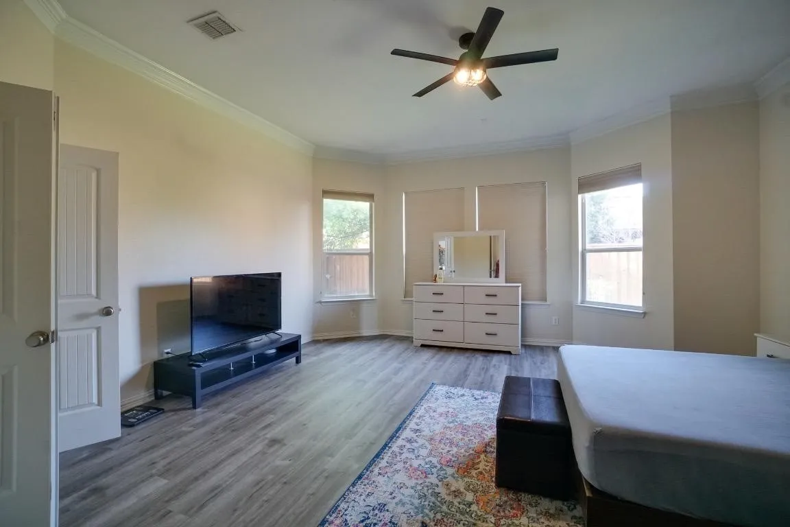 Bay window with multiple windows, wood-finish flooring, ceiling fan with integrated lighting, crown molding, and neutral wall tones