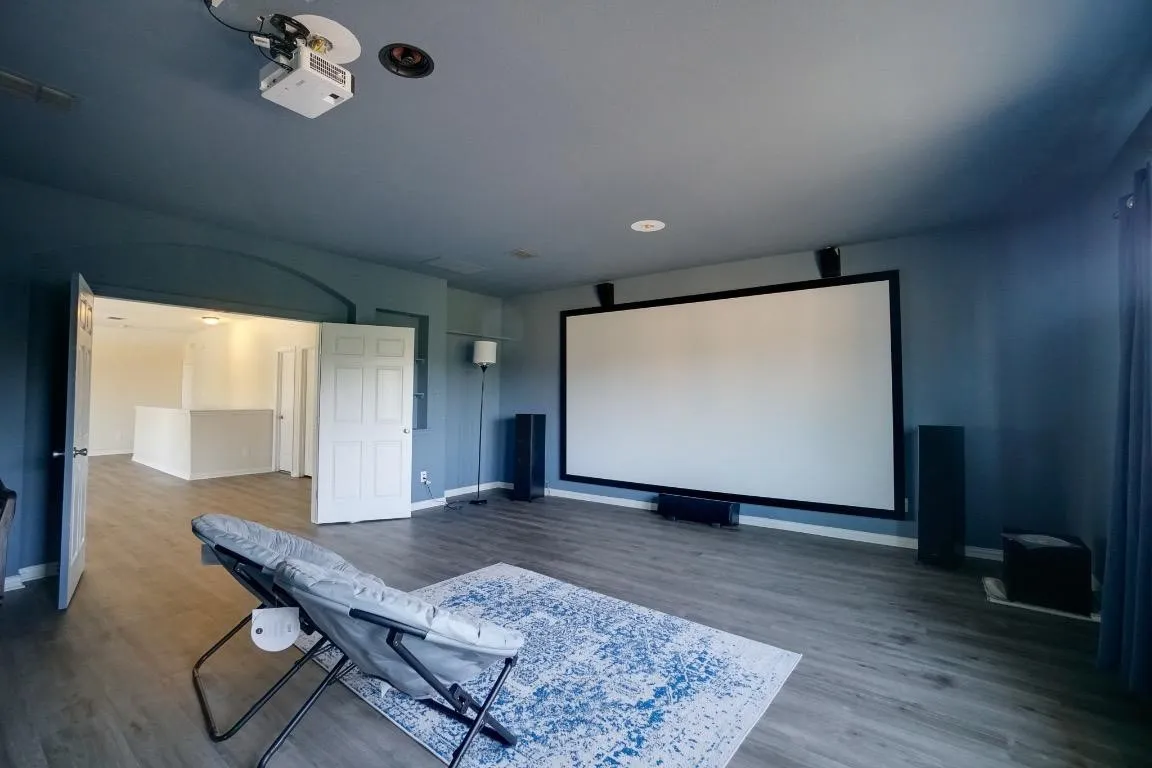 Large projection screen wall with ceiling-mounted projector, recessed lighting, wood-finish flooring, and arched doorway