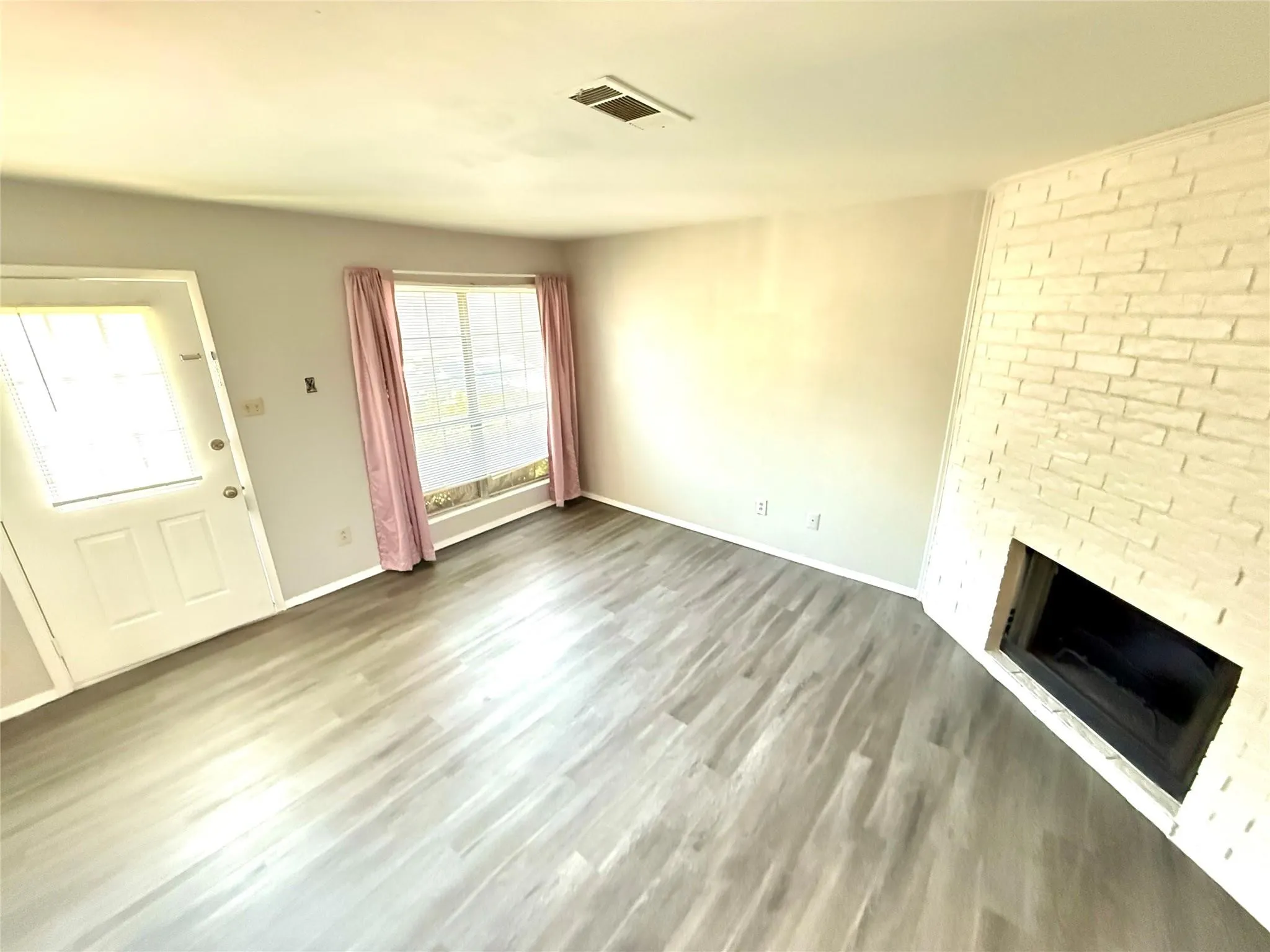 Living area featuring light wood-finish flooring, a white brick fireplace, and a large window with blinds