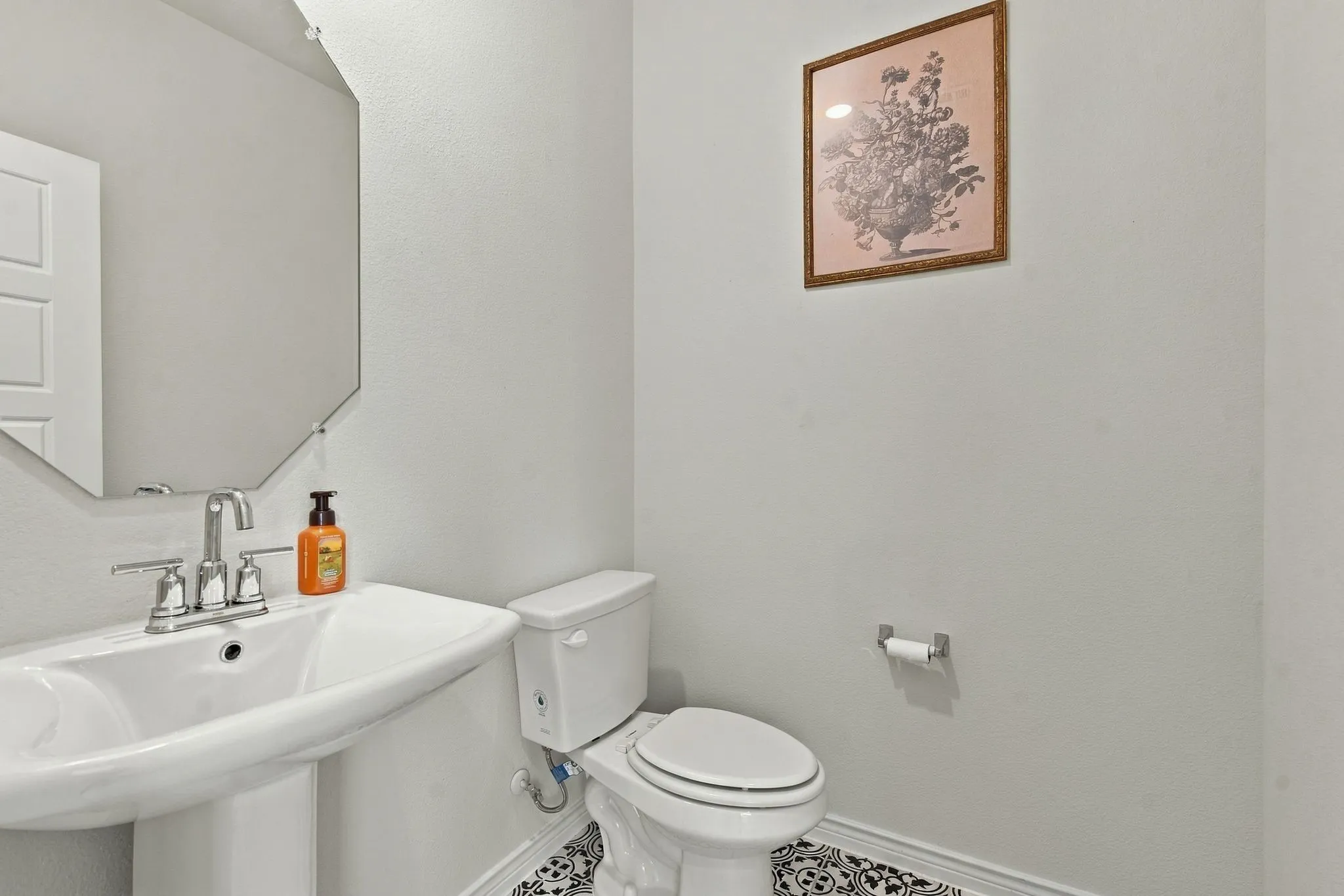 Pedestal sink with chrome faucet, octagonal wall mirror, toilet, and patterned tile flooring