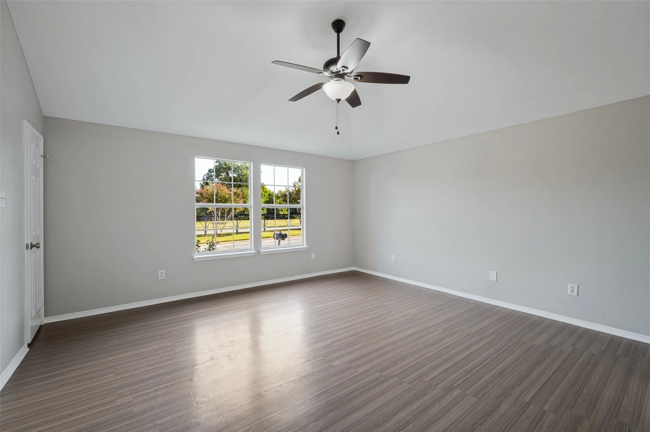 Spacious room featuring wood-finish flooring, light grey wall paint, a ceiling fan with integrated lighting, and two large windows providing natural illumination
