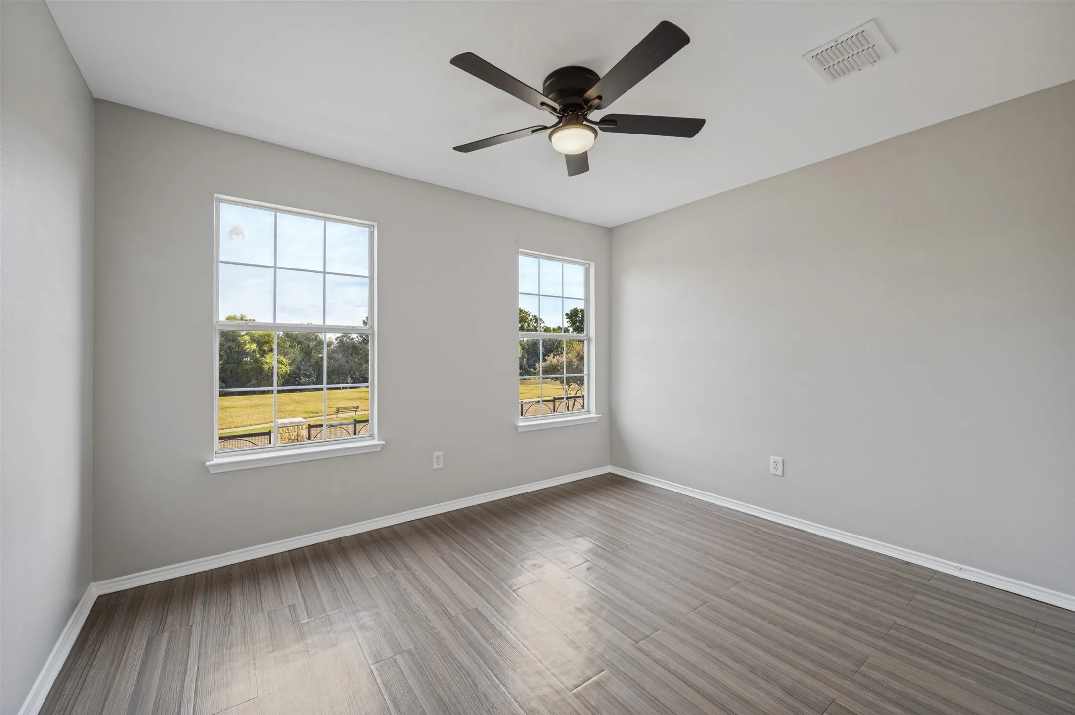 Spacious room featuring wood-finish flooring, light gray wall paint, two large windows, a ceiling fan with integrated lighting, and white trim throughout