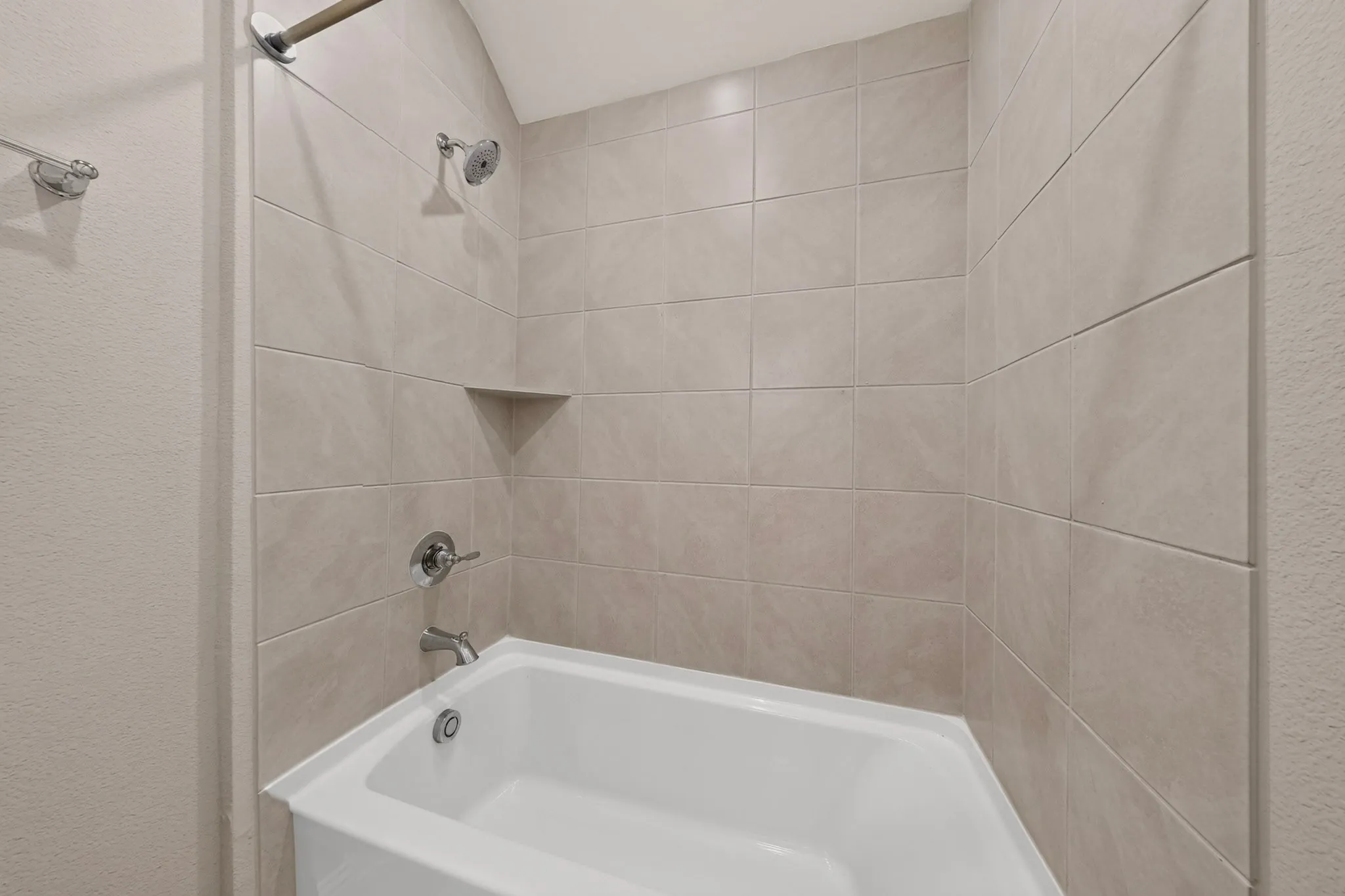 Built-in bathtub with neutral-toned wall tiling, chrome fixtures, and a recessed shower shelf