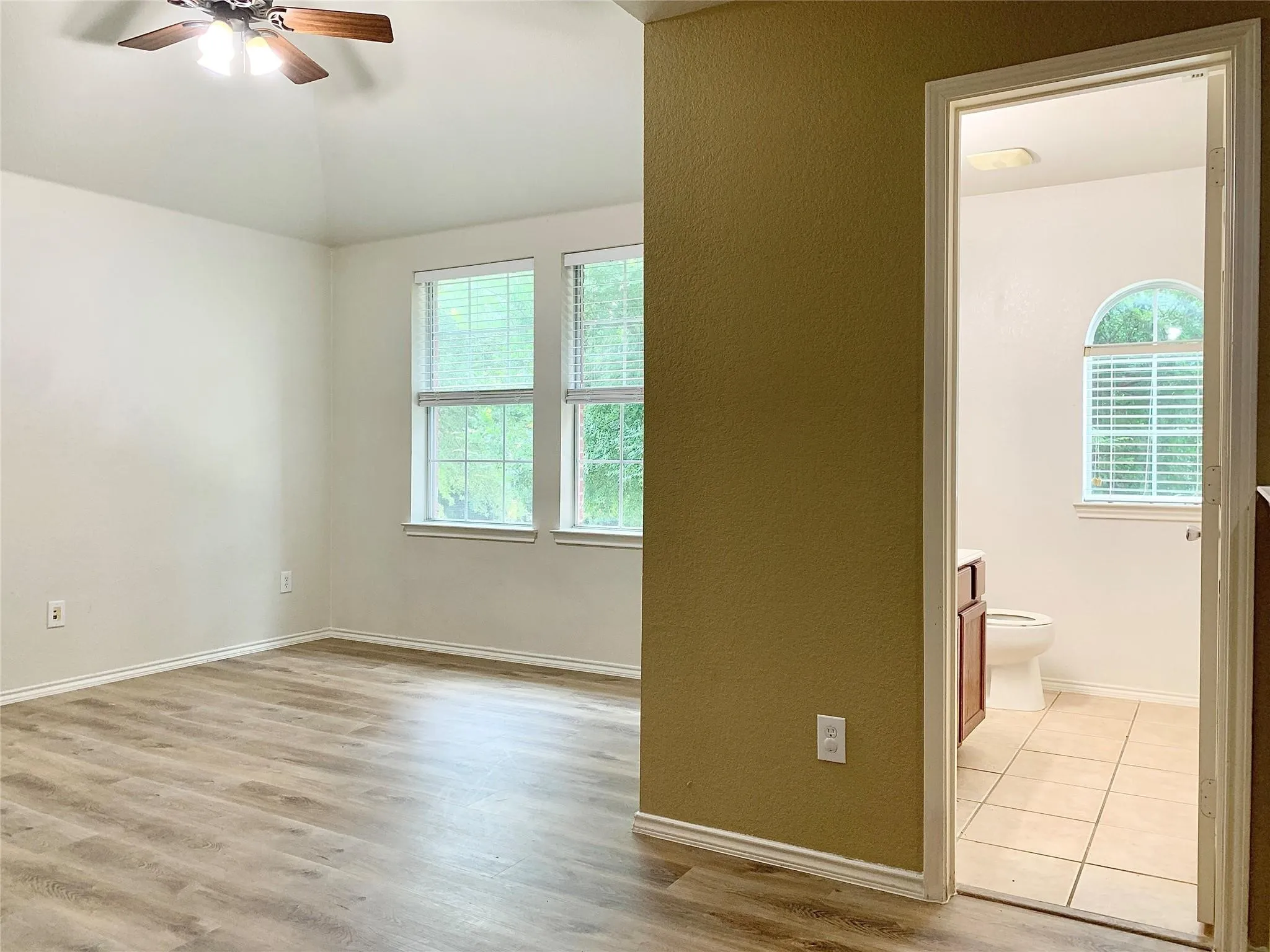 Wood-finish flooring, ceiling fan with integrated lighting, multiple windows with blinds, and an arched window in the bathroom
