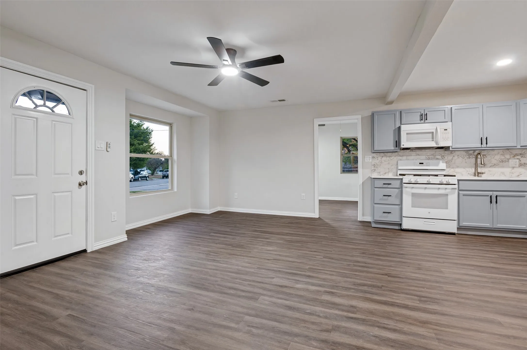 Wood-finish flooring throughout, modern ceiling fan with integrated lighting, white appliances, grey cabinetry, and a decorative front entry door