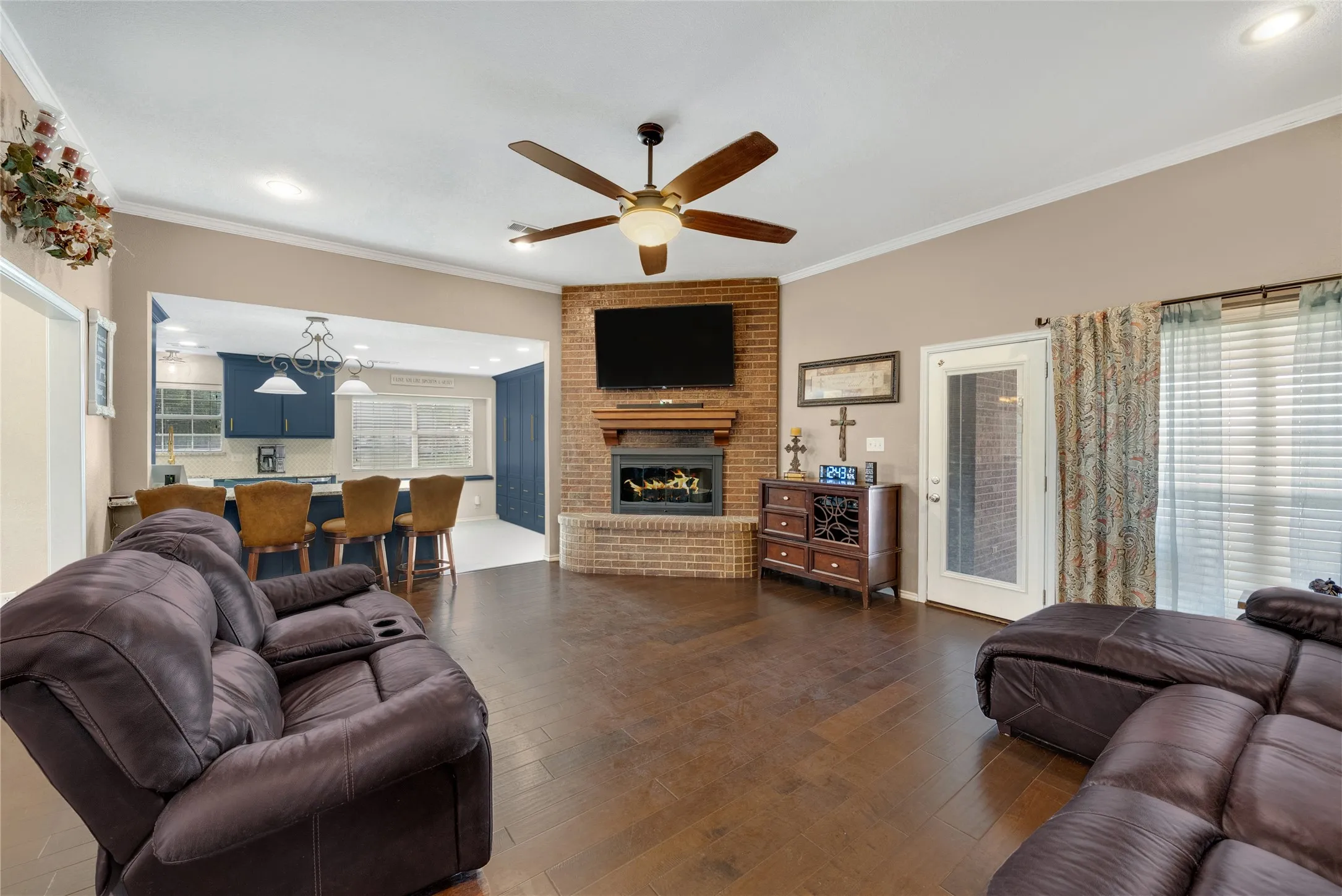 Spacious living area featuring wood-finish flooring, a brick fireplace with a wood mantel, and a ceiling fan with integrated lighting