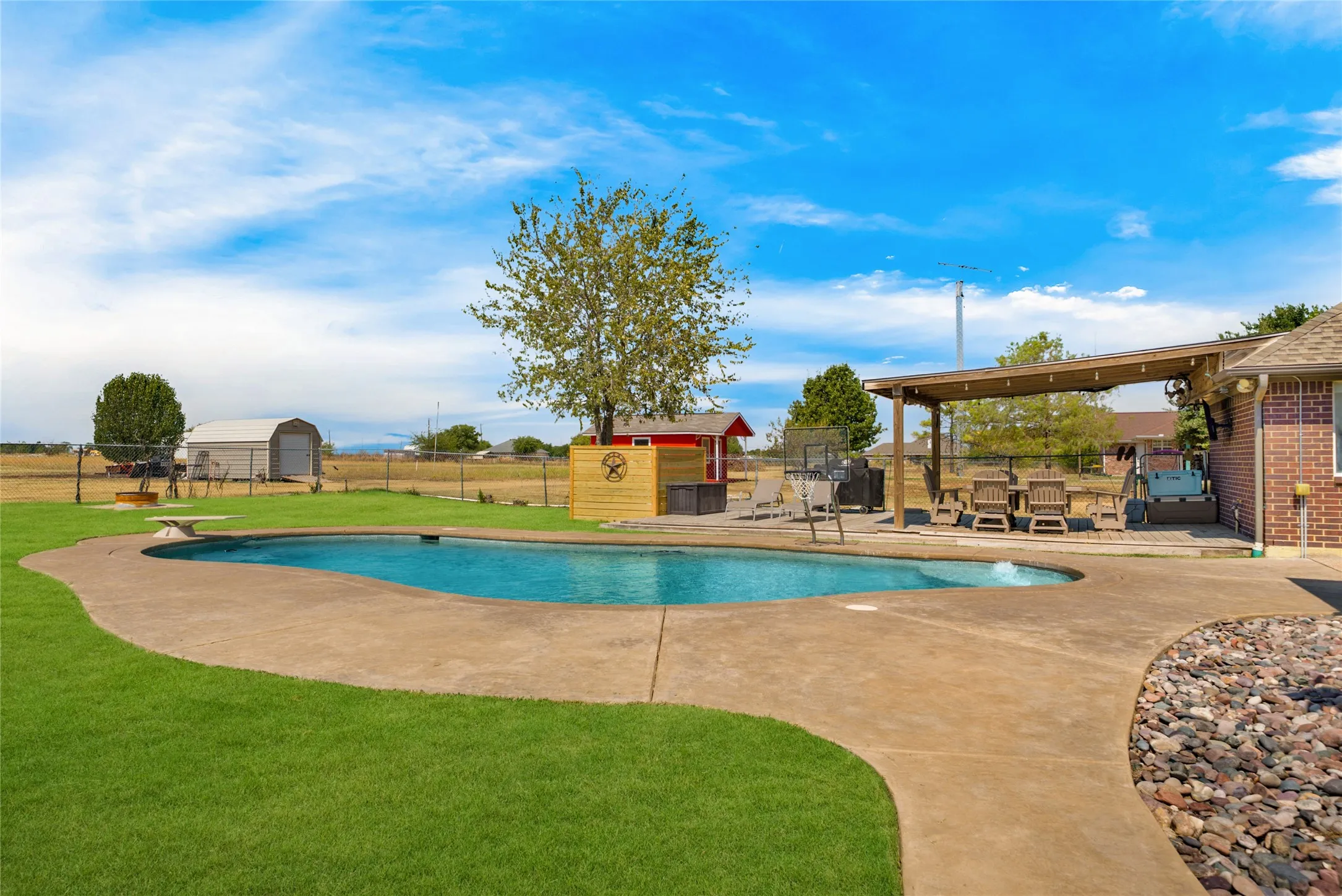 Curved swimming pool with concrete decking, a covered patio structure, and a brick exterior wall