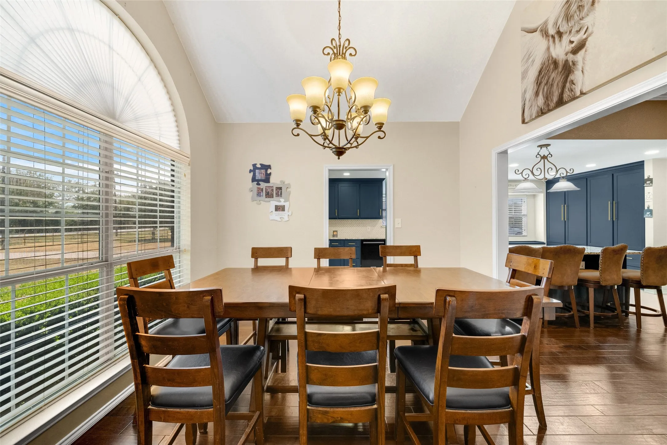 Vaulted ceiling dining area with wood-finish flooring, featuring a large arched window with horizontal blinds and an ornate chandelier