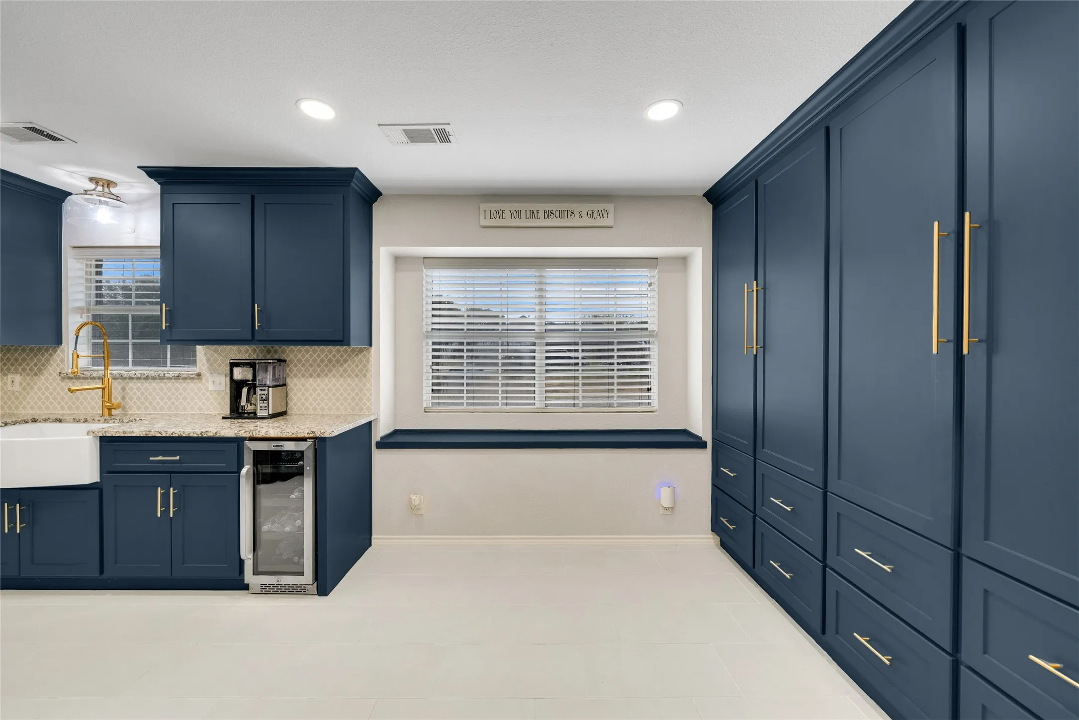Navy blue cabinetry with gold-tone hardware, built-in beverage cooler, farmhouse sink, mosaic tile backsplash, and light-tone tile flooring