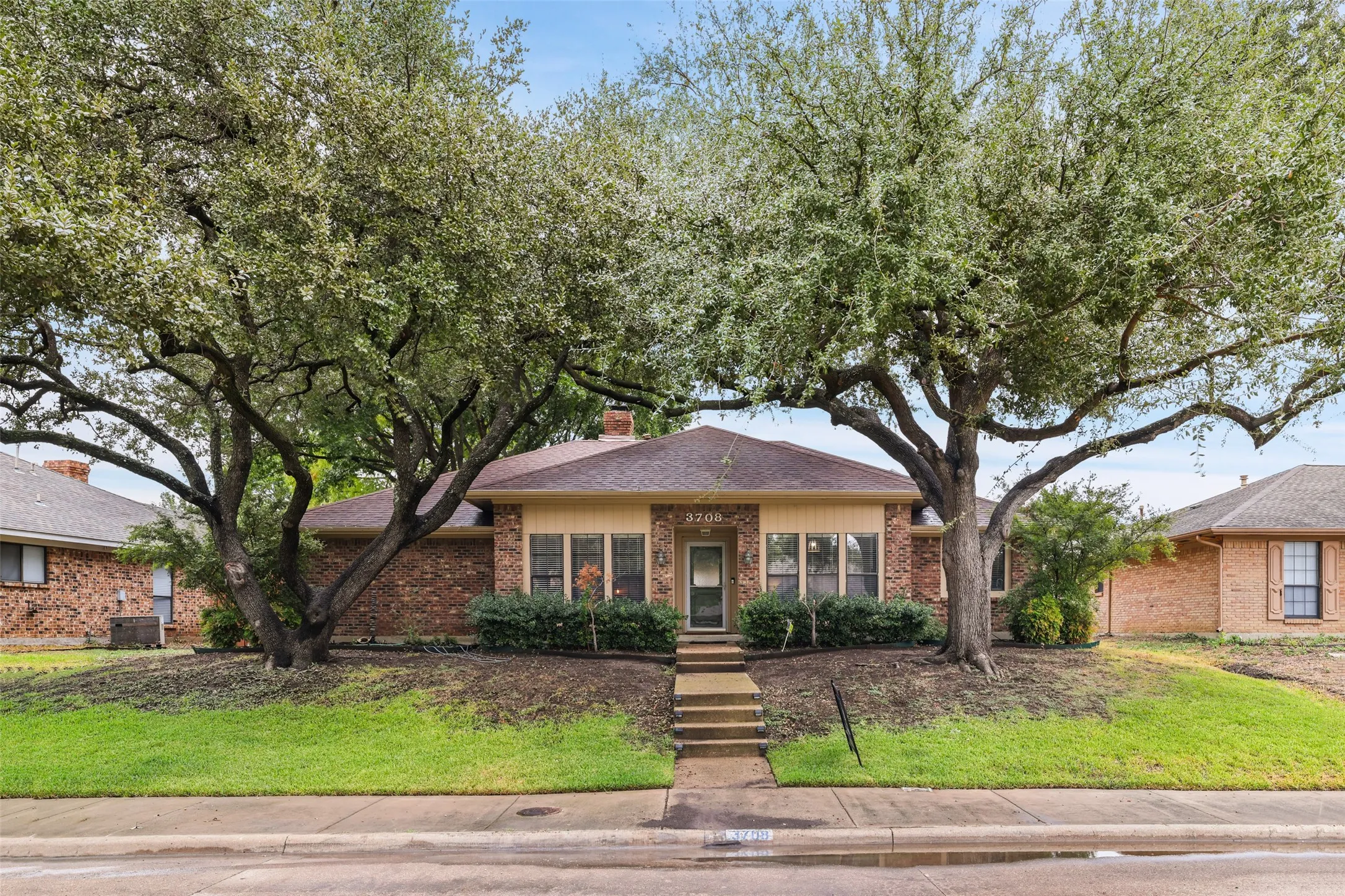 Traditional brick exterior with a brown shingle roof, mature front yard trees, and a concrete pathway leading to the entry