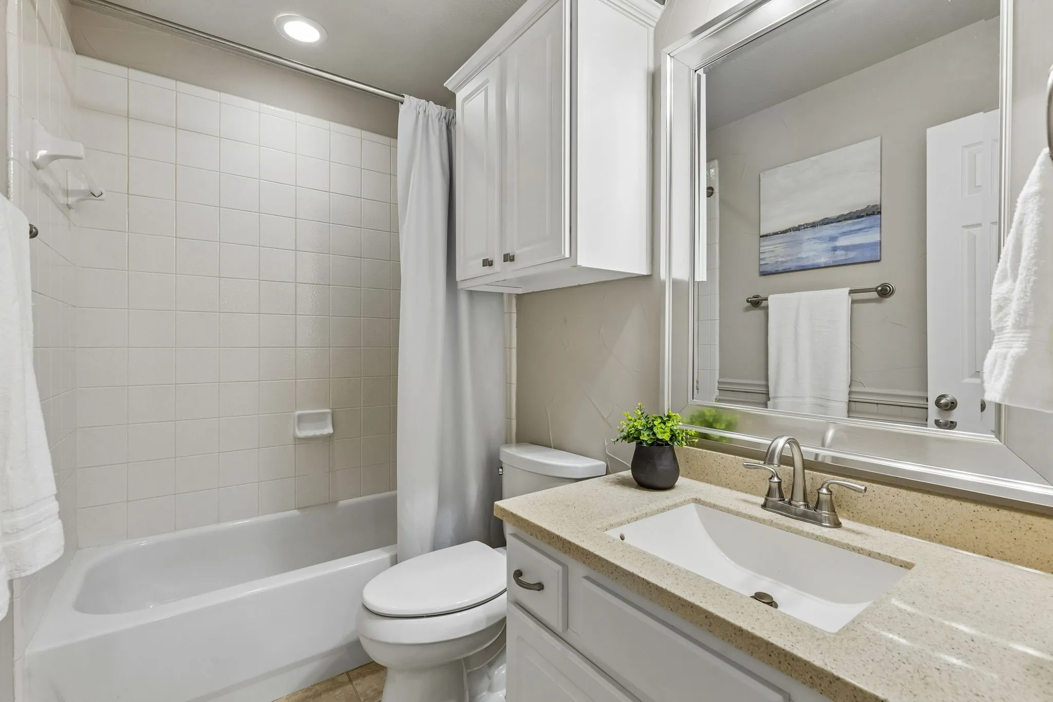 White tile tub/shower combination, white toilet, white vanity with integrated sink, light-colored speckled countertop, and wall-mounted mirror