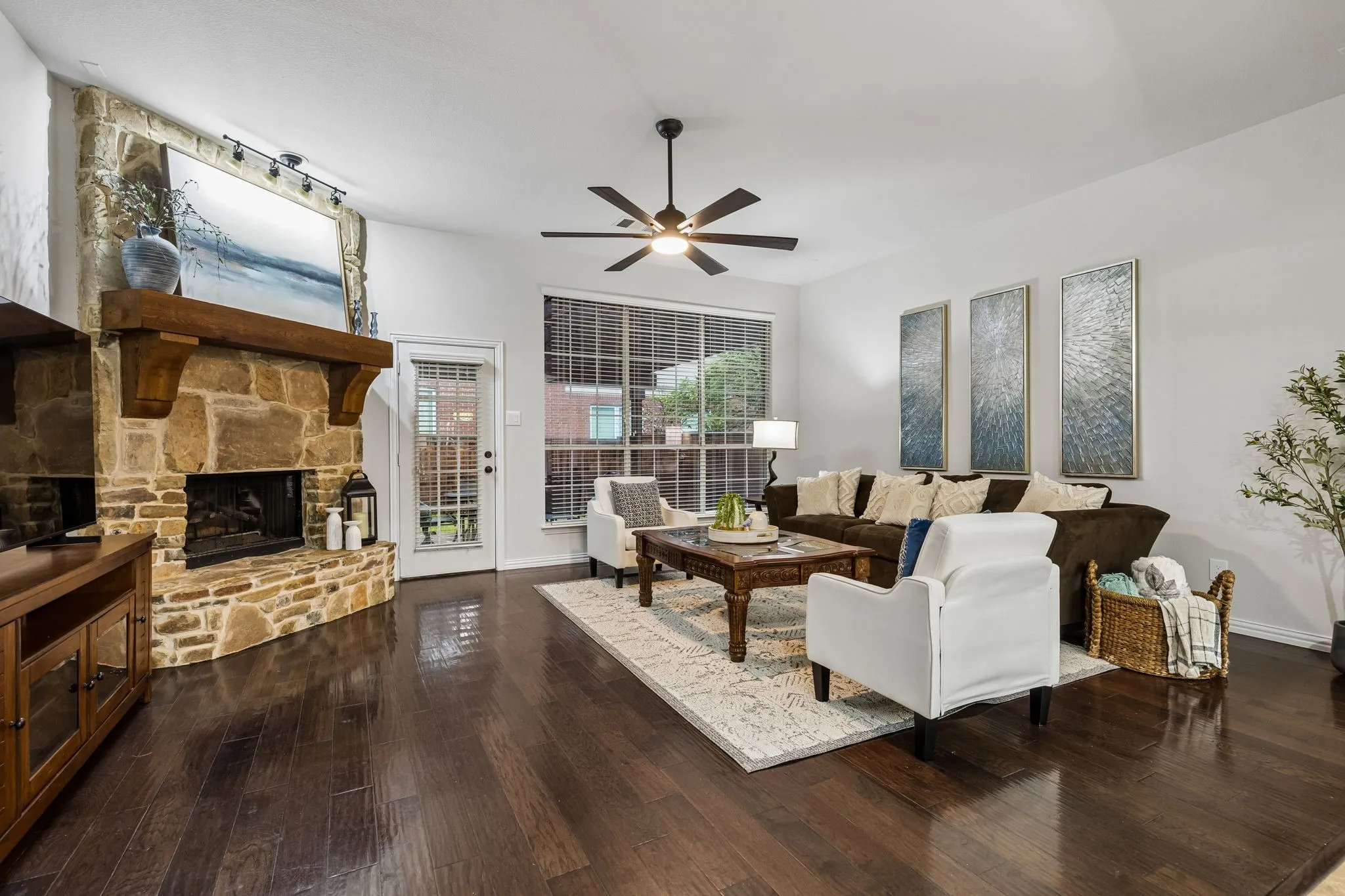 Hardwood-finish flooring, stone-clad fireplace with wood mantel, large windows with blinds, ceiling fan, and white walls
