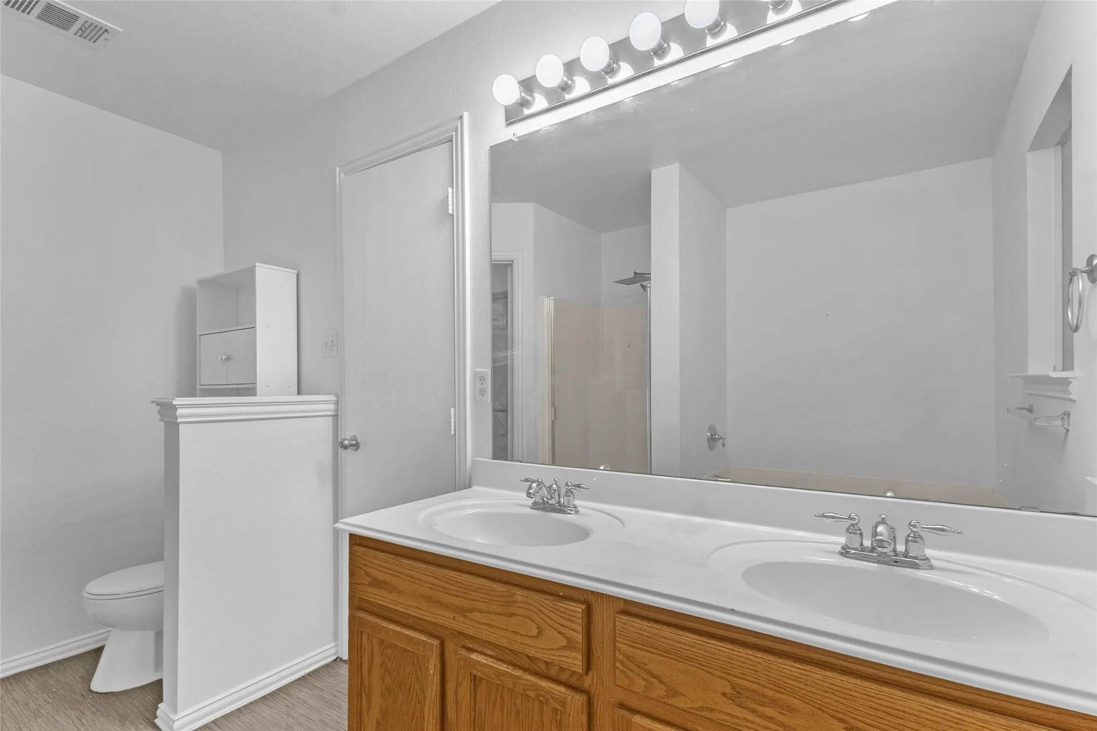 Primary bath double vanity with wood-tone cabinets and a half-wall toilet nook