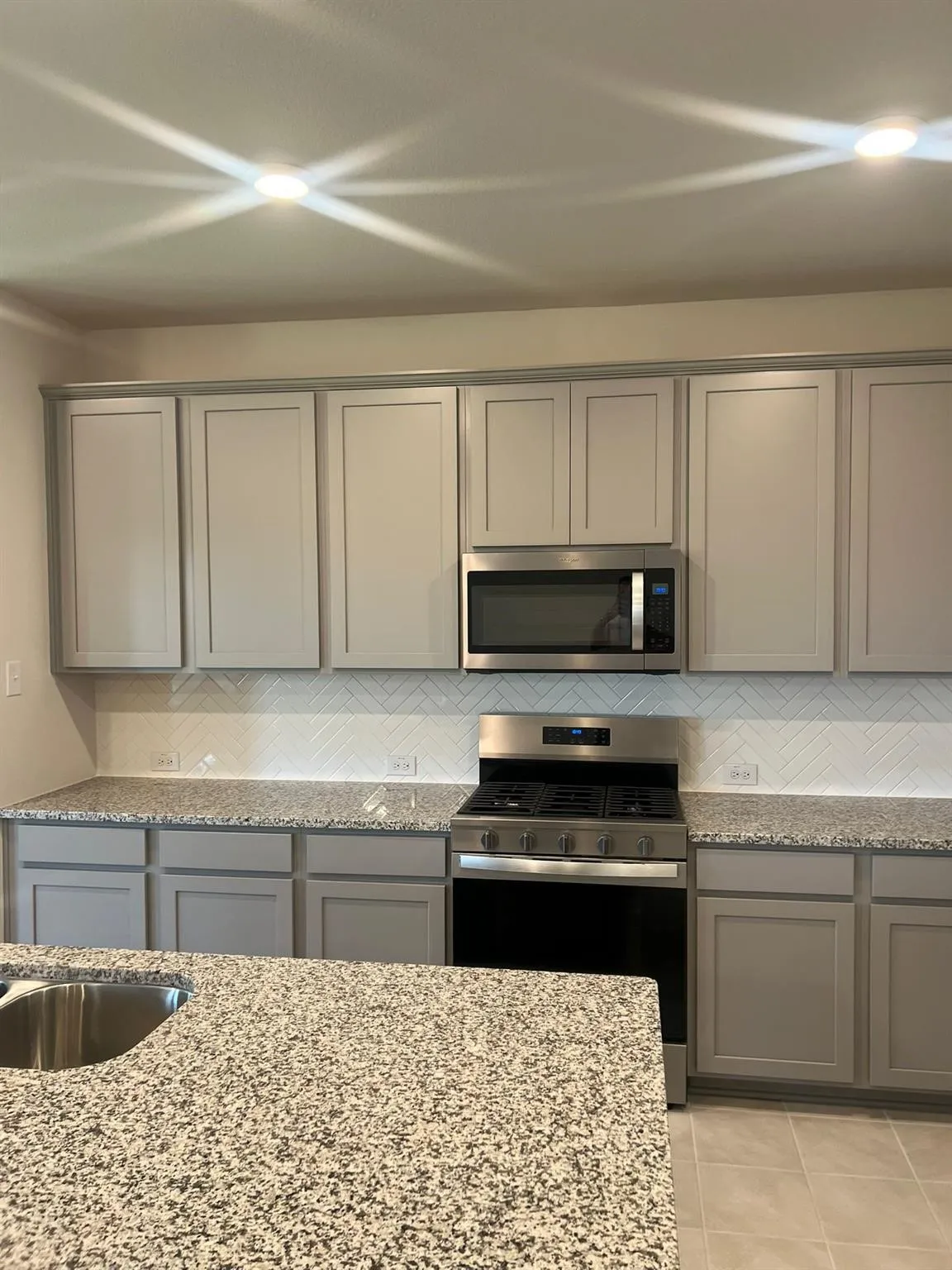 Kitchen featuring light gray cabinetry, stainless steel appliances, granite countertops, and a herringbone tile backsplash